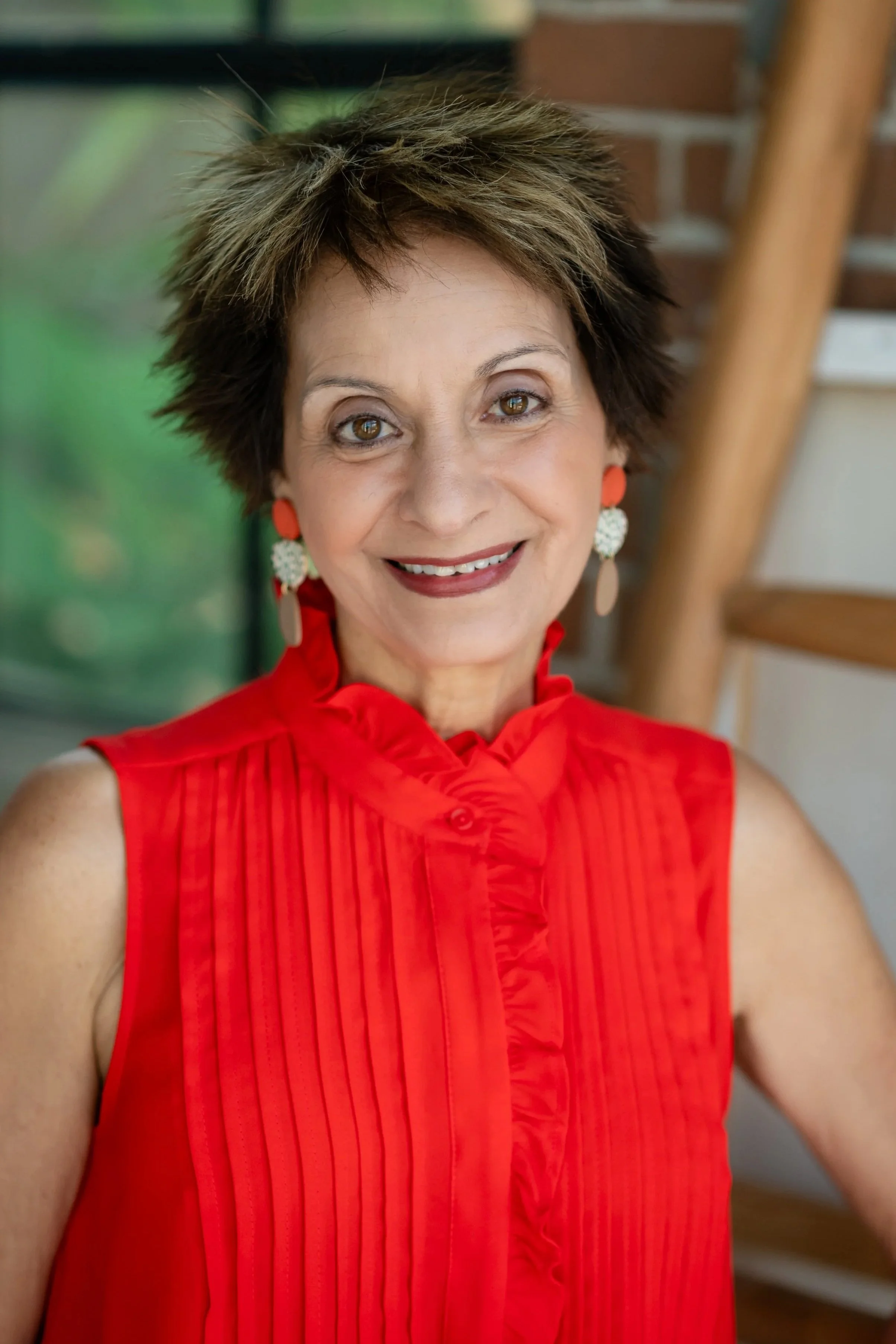 A woman with short, spiky hair wearing a bright red sleeveless blouse and large earrings, smiling at the camera in an indoor setting.