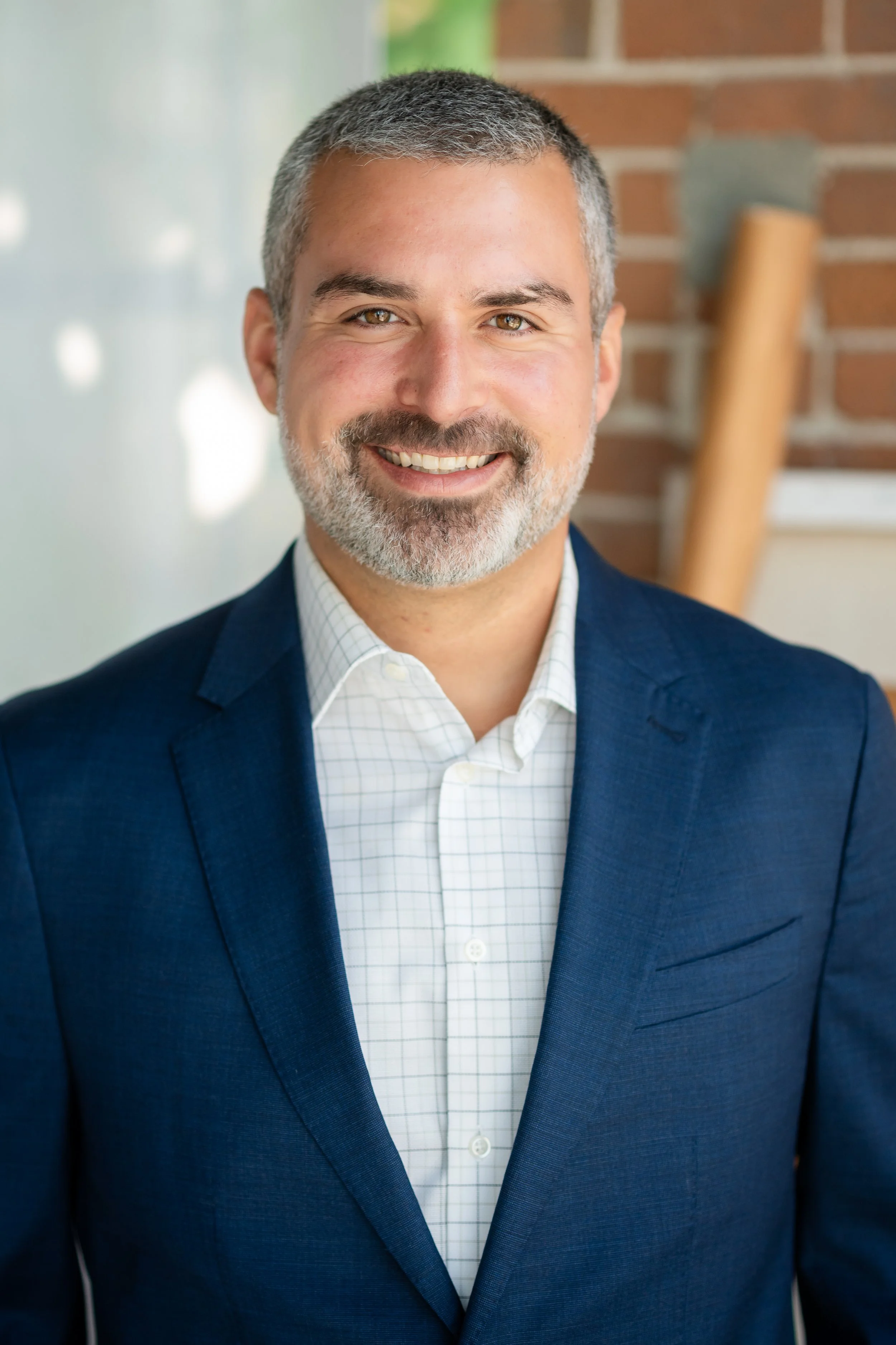 Headshot of a smiling man with short gray hair and beard, wearing a navy blazer and white checkered shirt, standing in front of a brick wall.