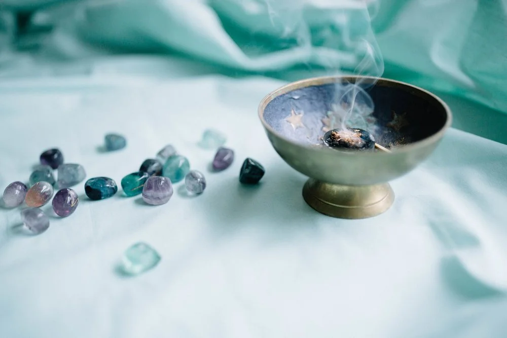 A brass incense holder with smoke rising from loose incense on a charcoal disk, surrounded by scattered small, crystal runes on a pastel green fabric surface.