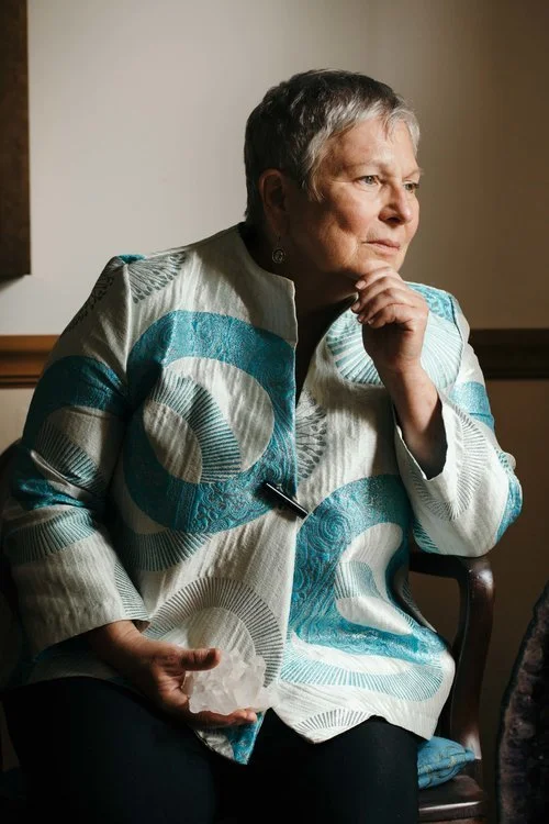 An older woman with short gray hair, wearing a patterned jacket in shades of blue and white, sitting indoors with her hand on her chin, looking thoughtfully out of the frame.