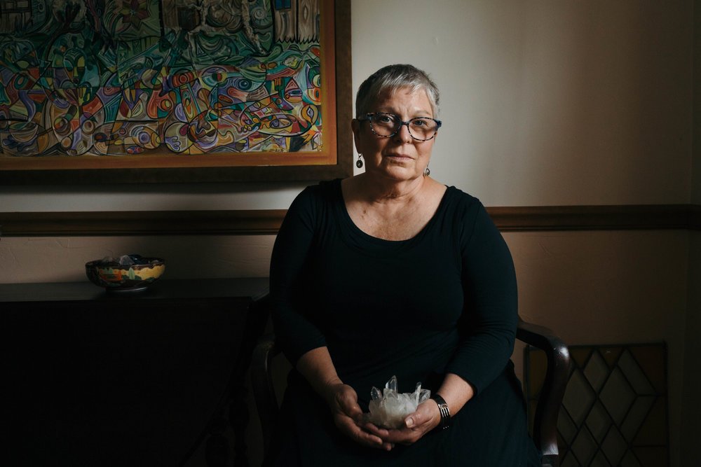 An older woman with short gray hair and glasses, wearing a black dress, sitting in a dimly lit room, holding a large clear quartz crystal, with a colorful abstract painting on the wall behind her.