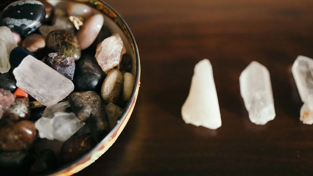 A bowl filled with various polished and raw crystals and stones on a dark wooden surface. Next to the bowl, there are three raw white quartz points arranged in a row.