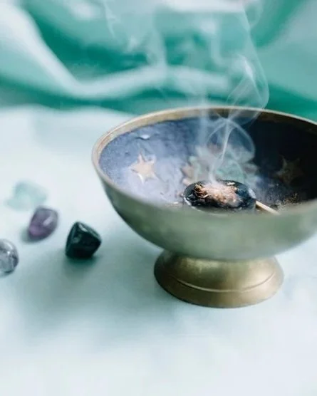 A burning incense cone in a brass incense holder shaped like a bowl with smoke rising from it, surrounded by small crystal runes on a light-colored surface.