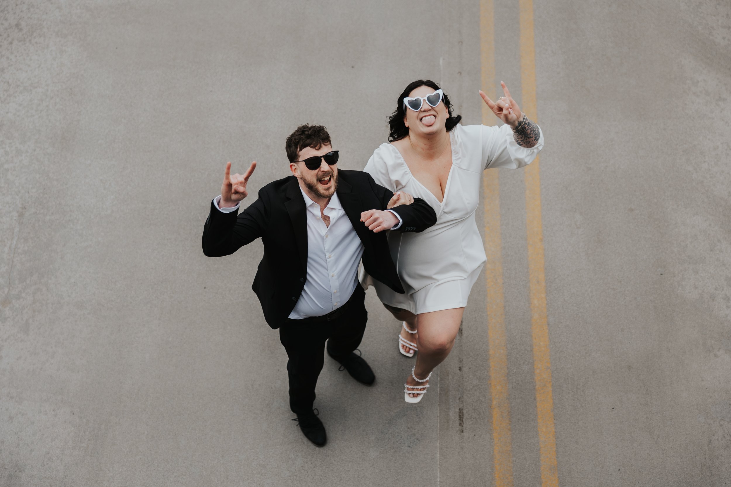 A man and woman dressed in formal attire celebrating and making rock and roll hand signs while standing on a street.