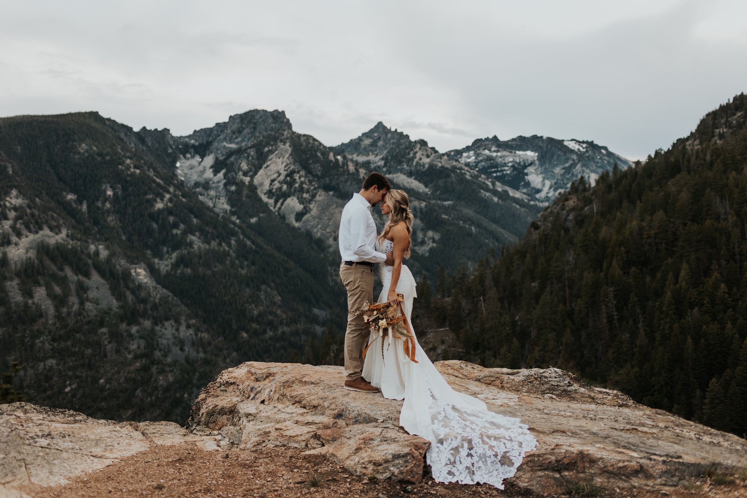 A couple dressed in wedding attire standing close together on a large rock in a mountainous landscape with trees and snow-capped peaks.