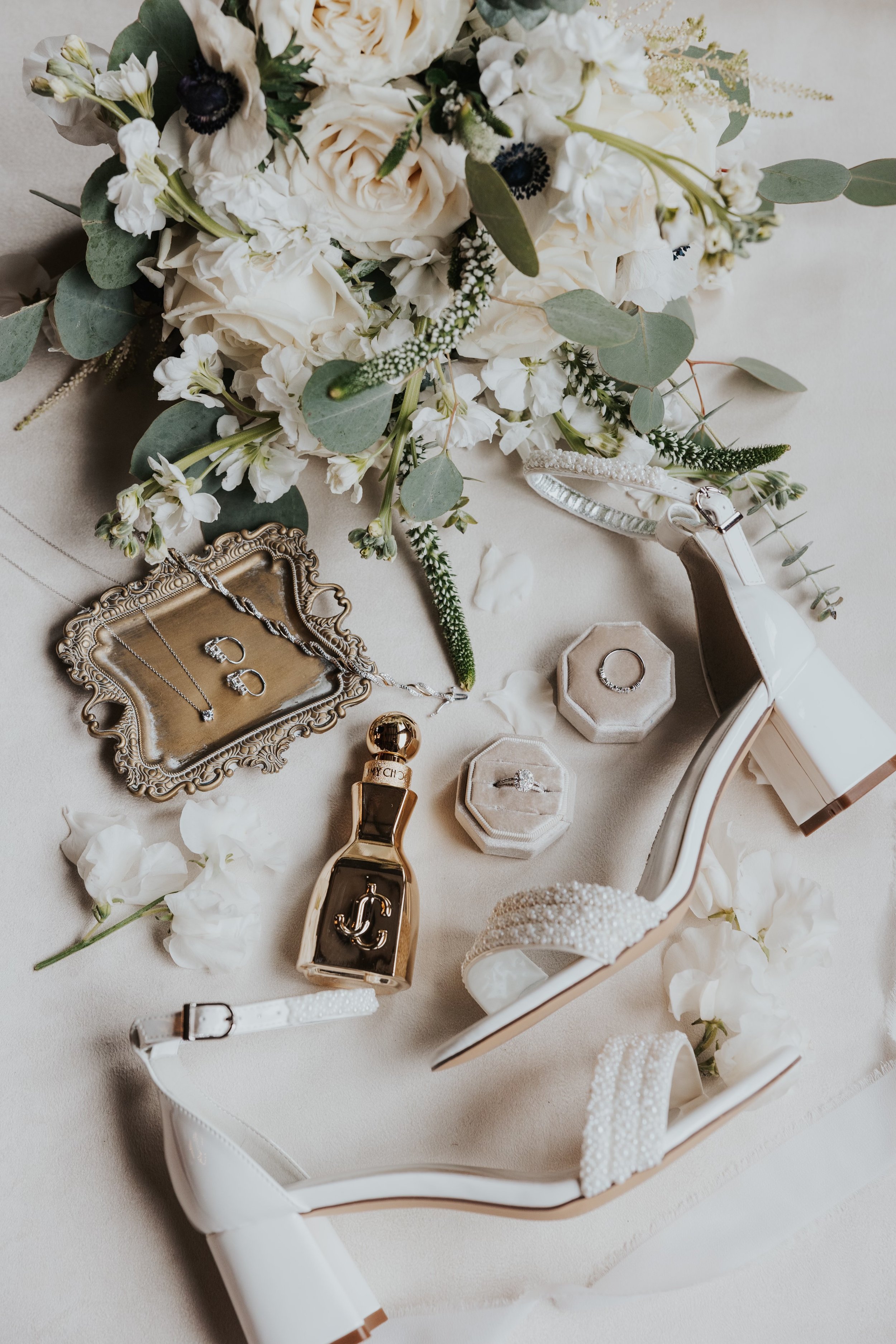 A collection of jewelry, a white high-heeled shoe, and a bouquet of white flowers with greenery on a light-colored surface.