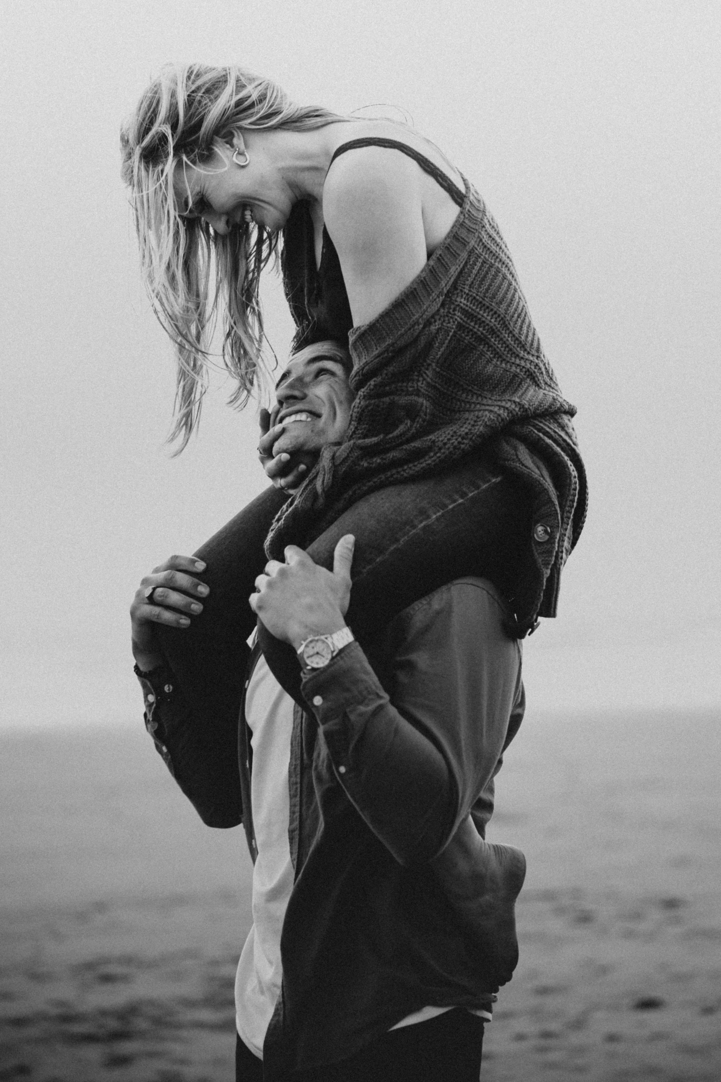 A black-and-white photo of a woman sitting on a man's shoulders, both smiling and looking at each other, outdoors near water.
