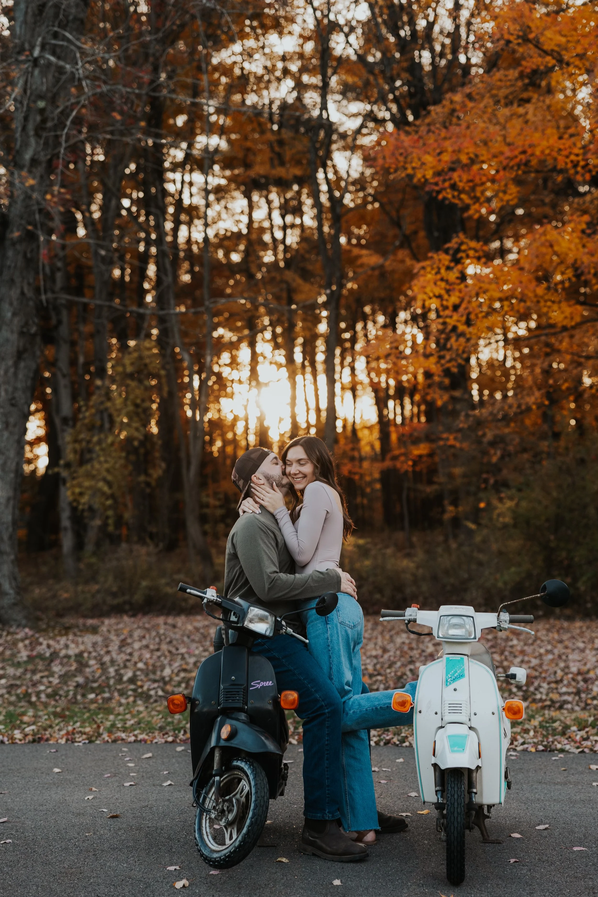 A couple sitting on scooters, embracing and smiling at each other during sunset in a forest with fall foliage.