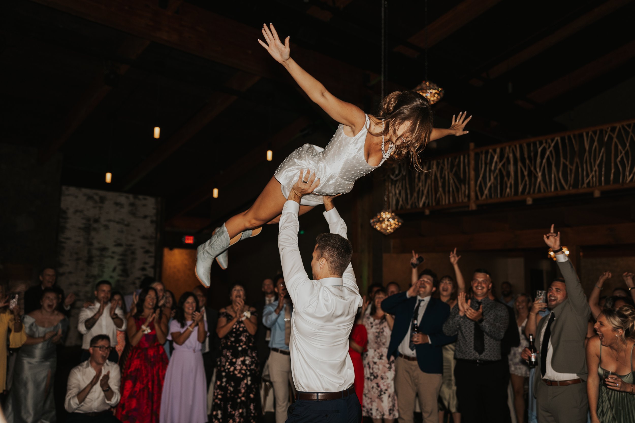 A bride being lifted in the air by a groom during her wedding reception, with guests cheering and clapping in the background.