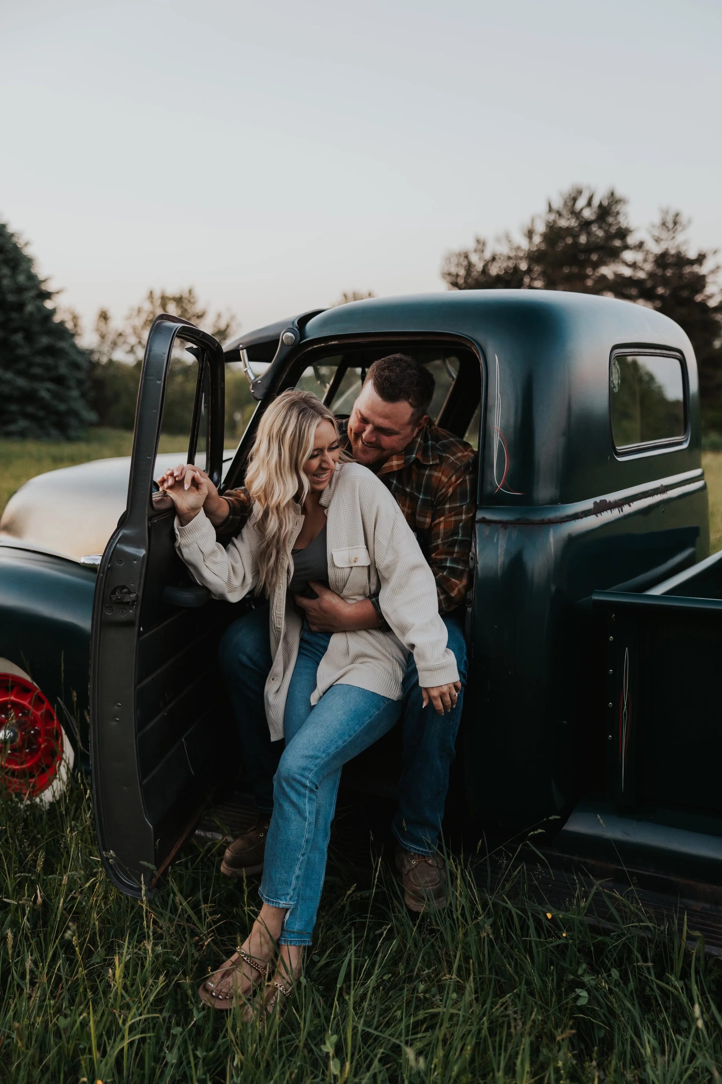 A smiling couple sharing a joyful moment near an old truck in a grassy field, with trees in the background
