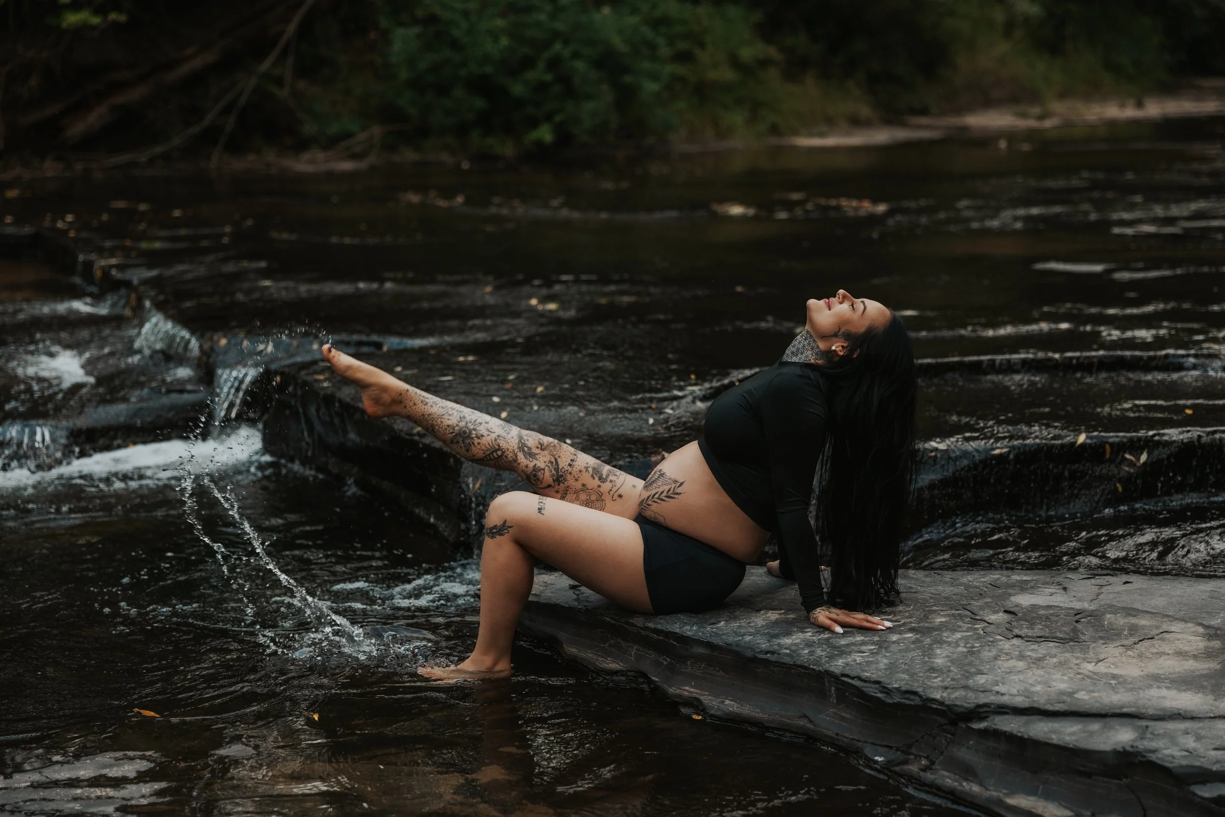 A woman with tattoos relaxing on a rock in a shallow river, leaning back with her eyes closed and long dark hair flowing down, wearing a black crop top and shorts.