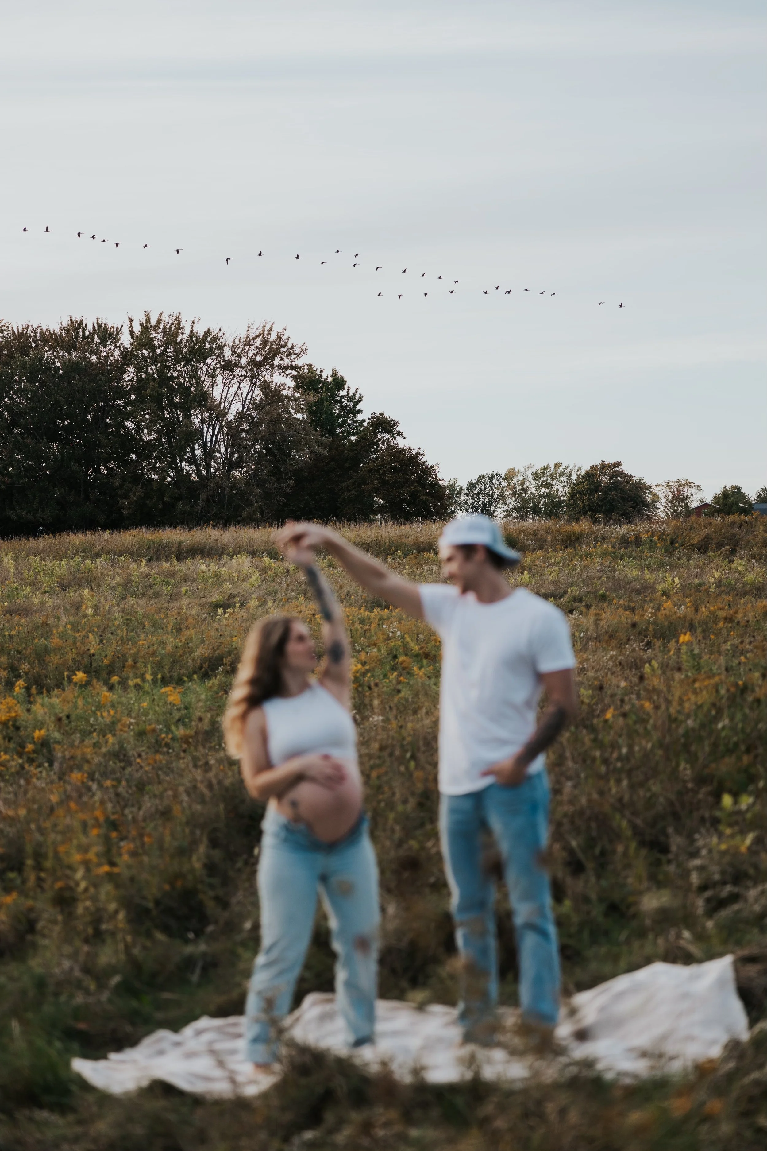 A pregnant woman and a man are standing outdoors in a field, holding hands, with the man lifting her hand. The woman is wearing a white tank top and ripped jeans, and the man is wearing a white t-shirt, light blue jeans, and a white cap. There are tr