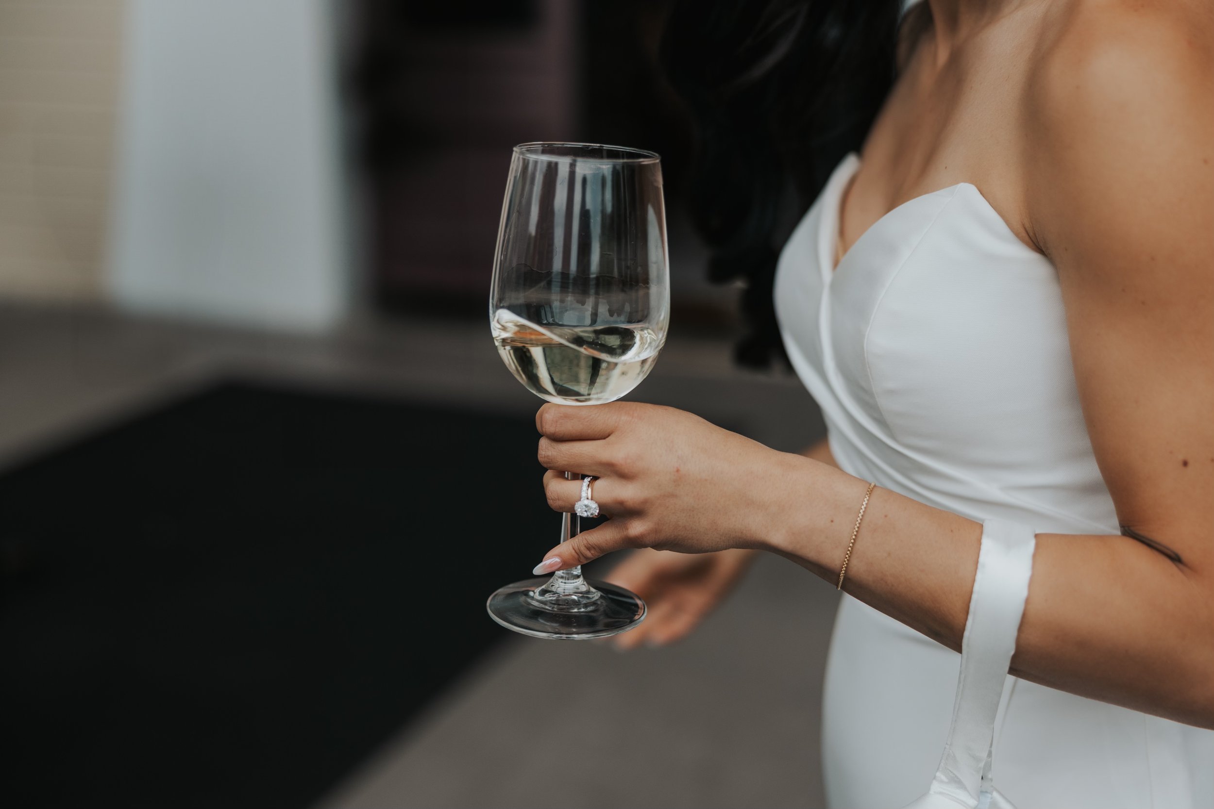 A woman in a white dress holding a glass of white wine with a diamond ring on her finger.