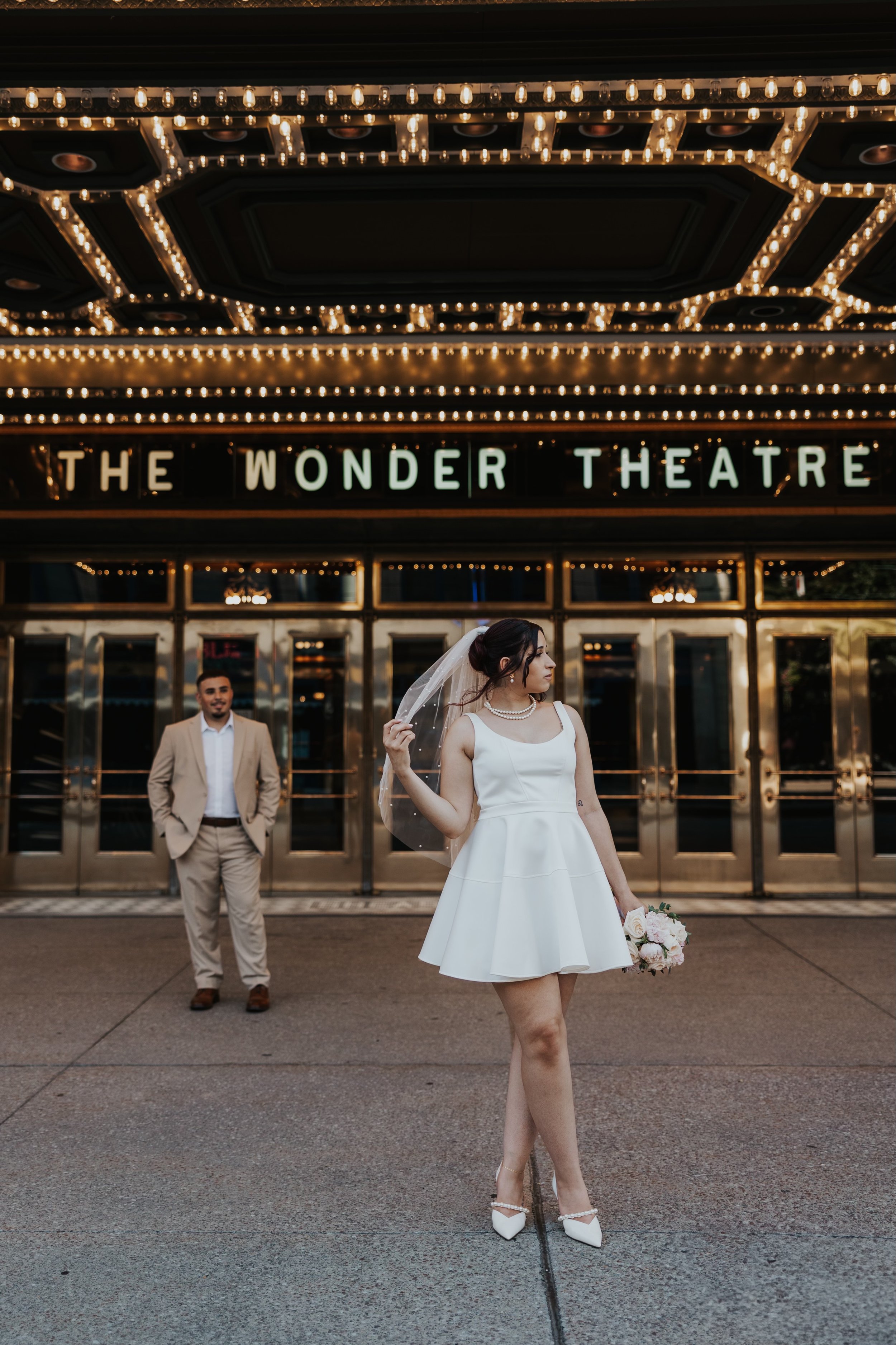 A bride and groom standing in front of The Wonder Theatre, with the bride holding a bouquet of flowers and wearing a white dress, while the groom stands behind her in a light-colored suit.