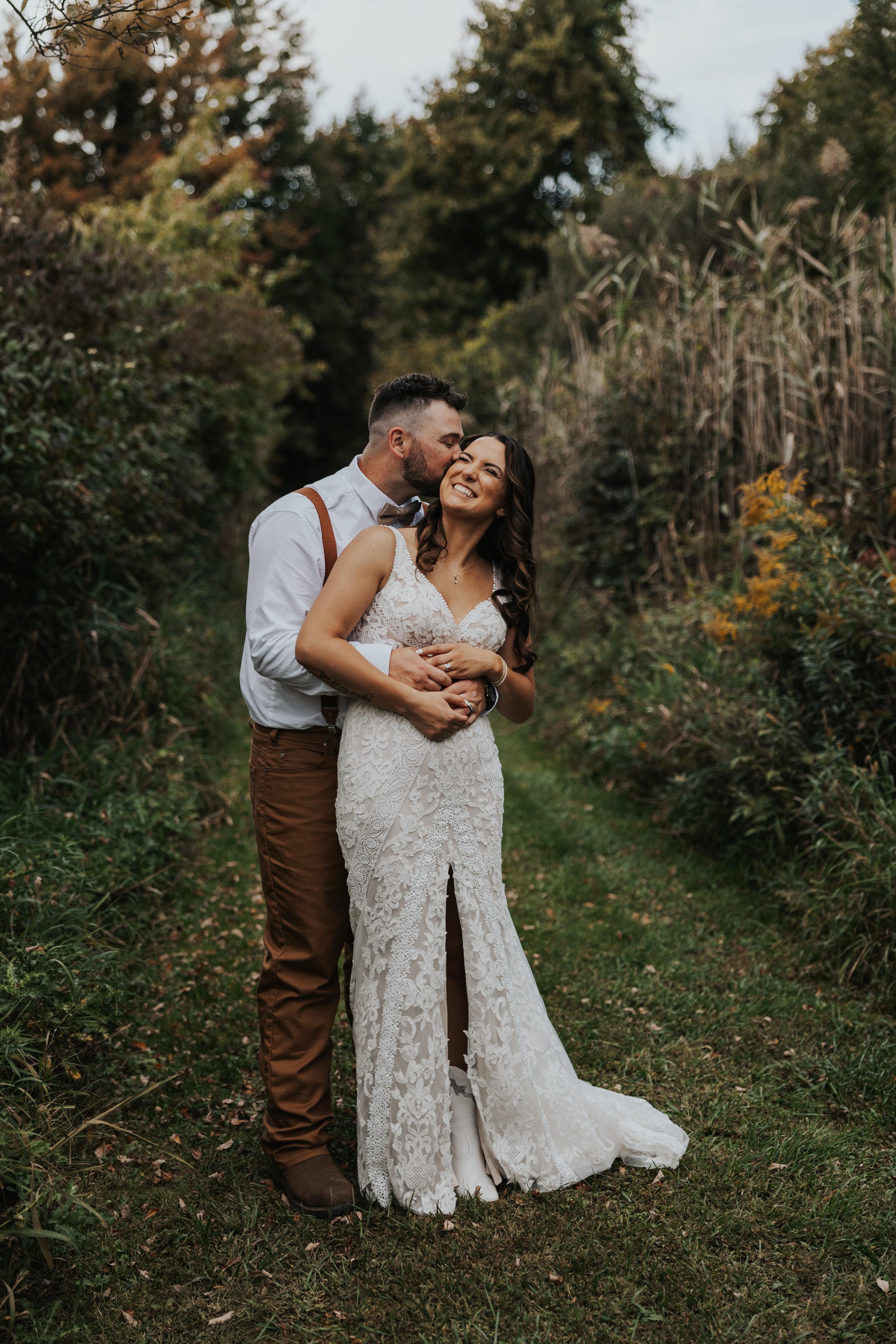 A happy couple in wedding attire standing on a grassy path surrounded by tall plants and trees, sharing an intimate moment in an outdoor setting.