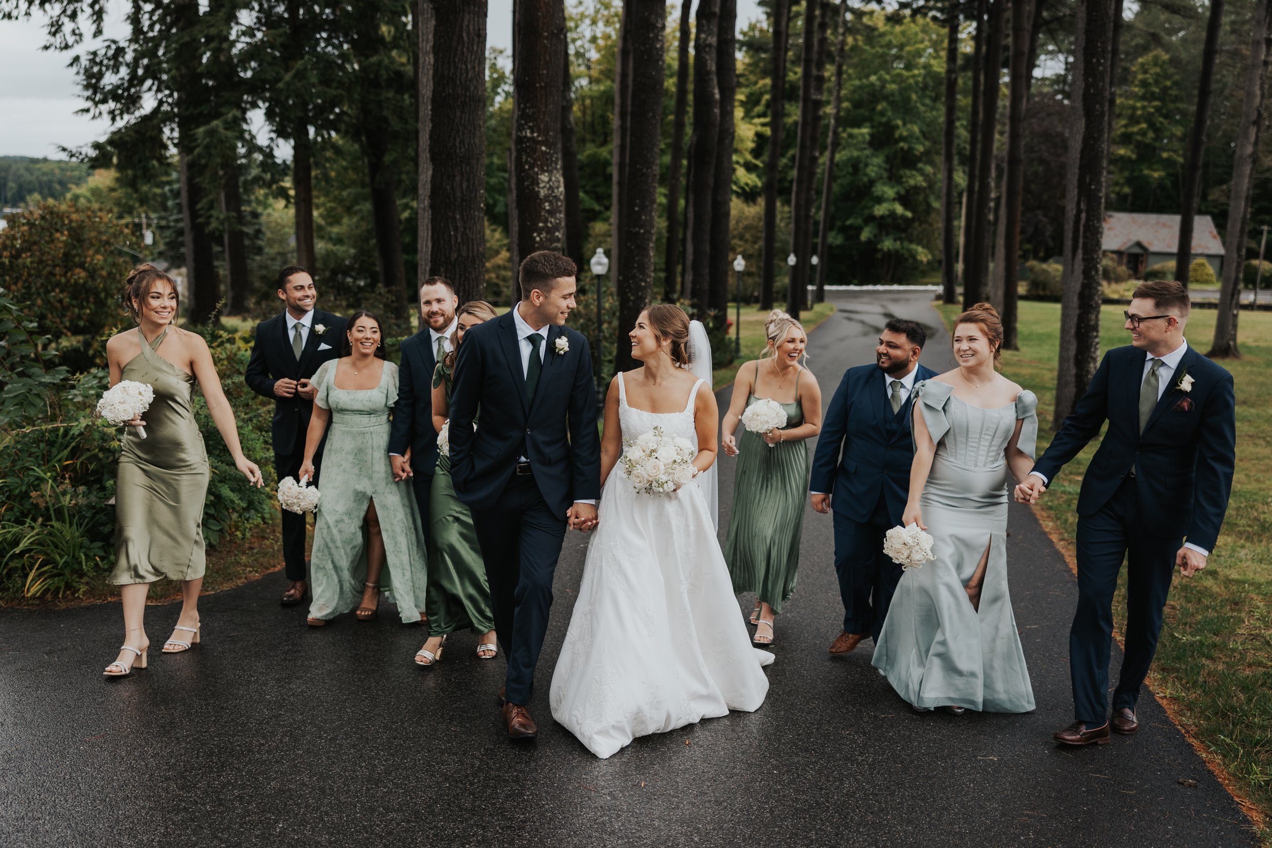A wedding party walking outdoors on a paved path through a wooded area, celebrating and smiling. The bride and groom walk hand in hand in the center, surrounded by bridesmaids and groomsmen dressed in elegant attire.