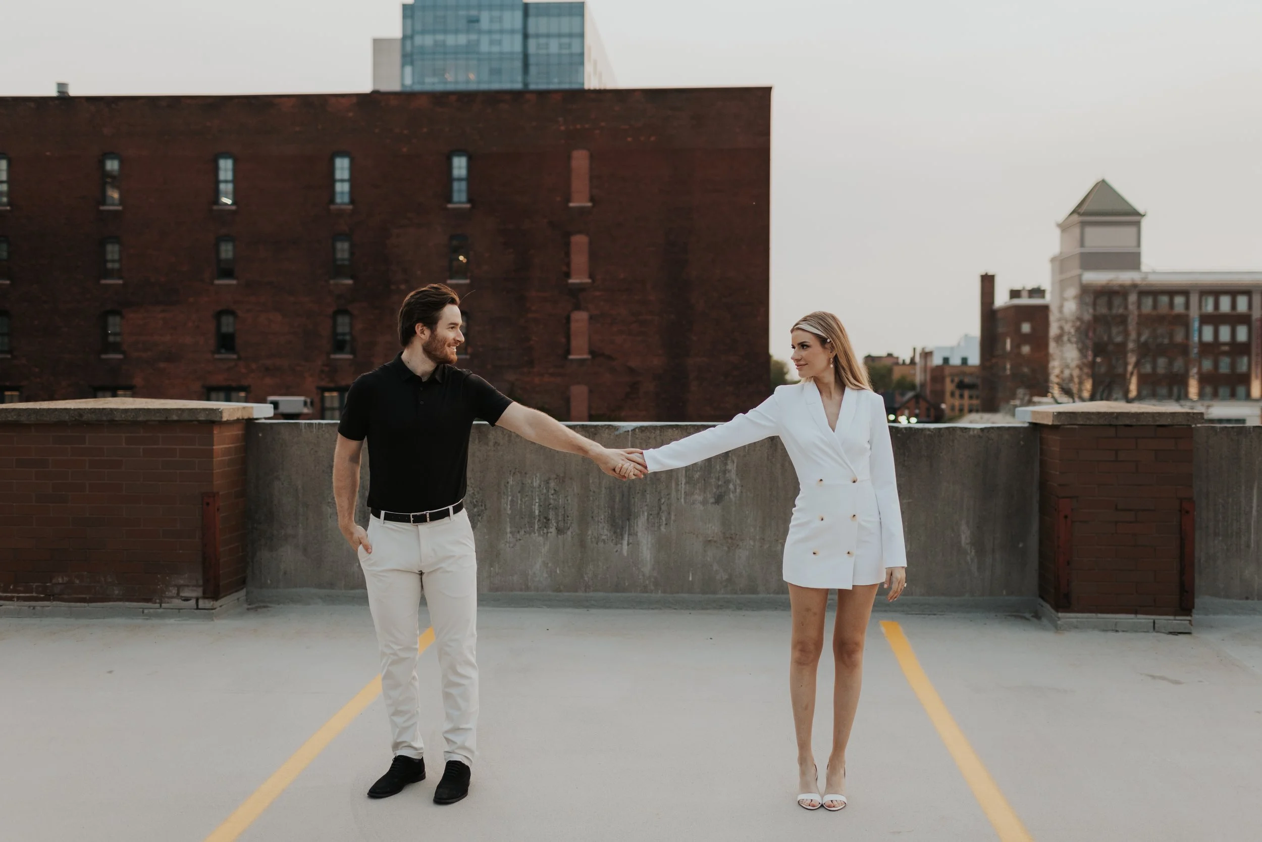 A man in a black shirt and white pants holding hands with a woman in a white blazer dress on a rooftop parking lot with urban buildings in the background.