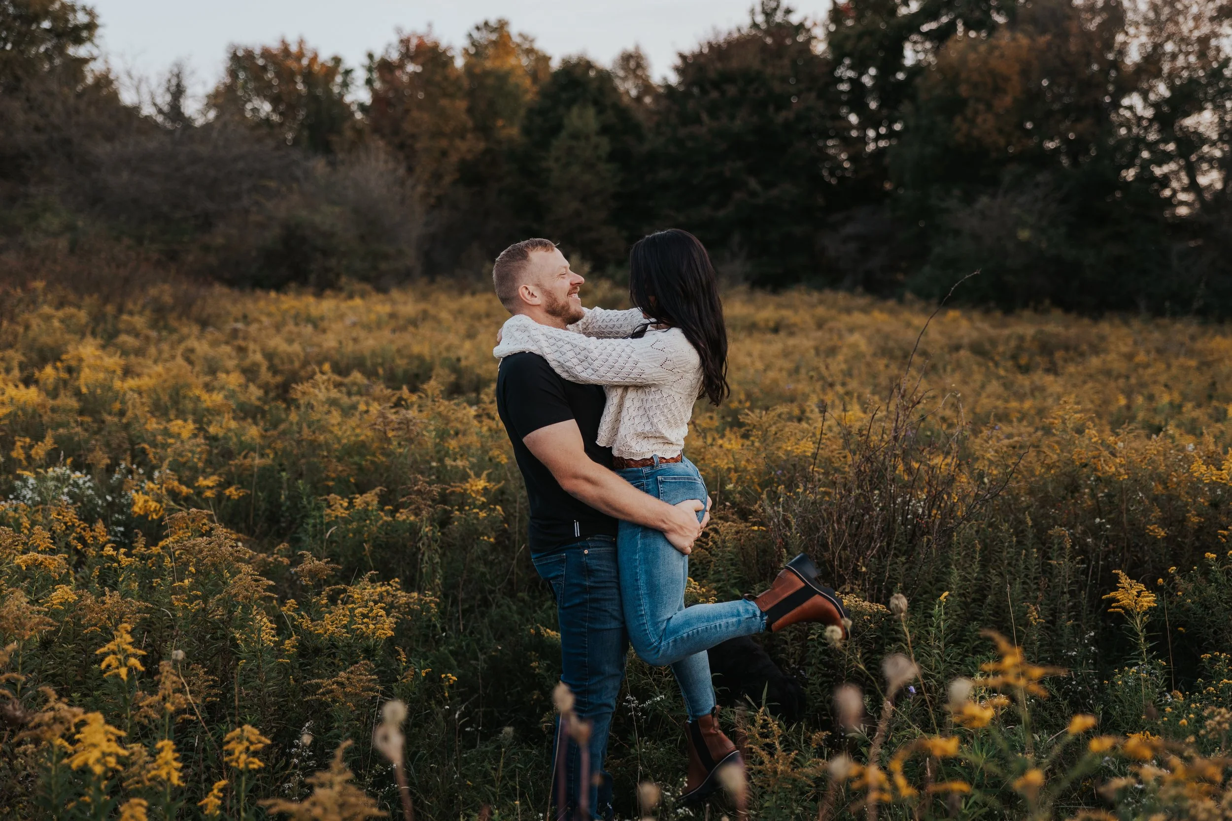 A couple embracing in a field of yellow flowers with trees in the background, during sunset.