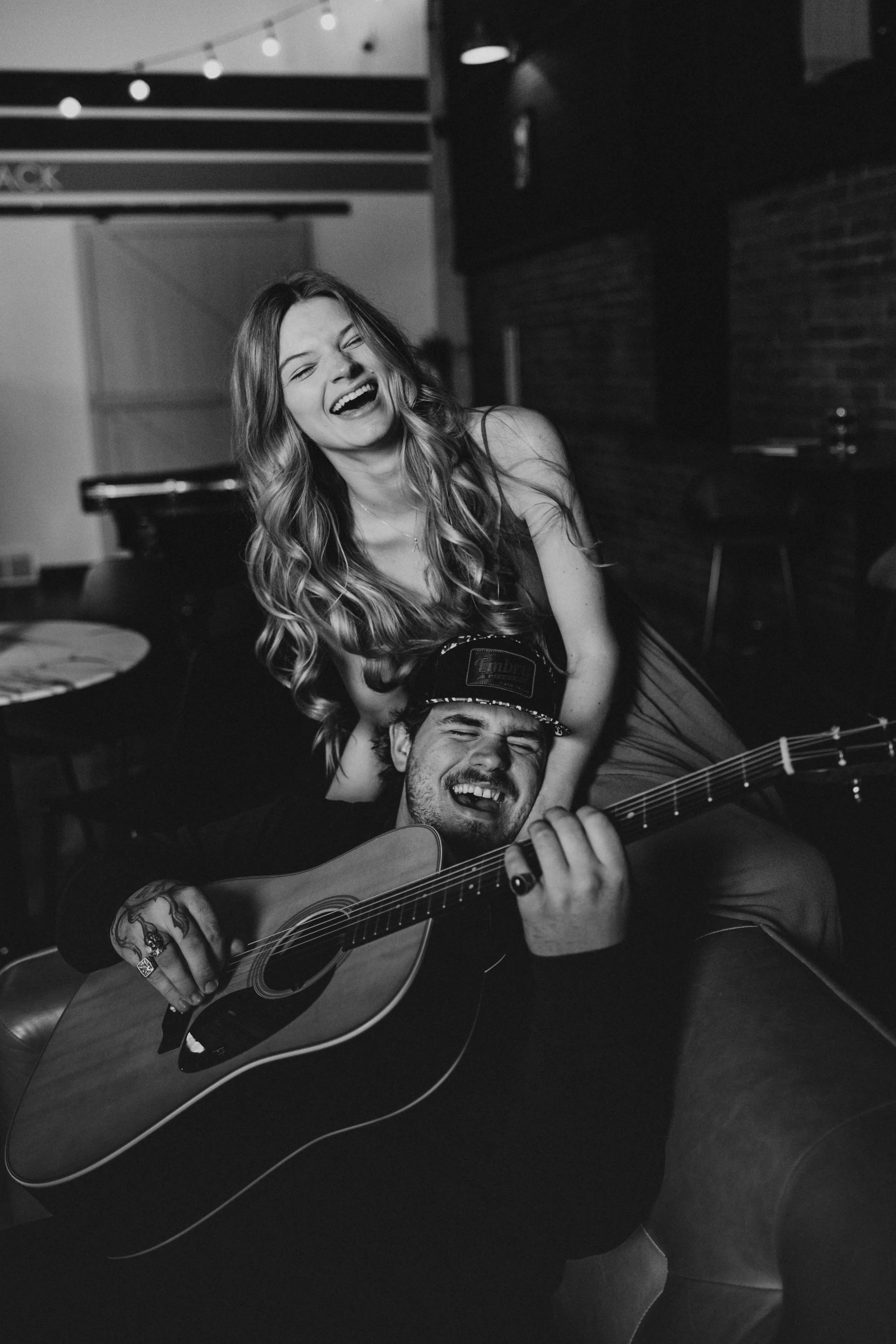 A black-and-white photo of a woman with long wavy hair laughing, sitting on a man's shoulders. The man is playing an acoustic guitar and wearing a baseball cap. They are in a dimly lit room with a brick wall and string lights visible in the backgroun