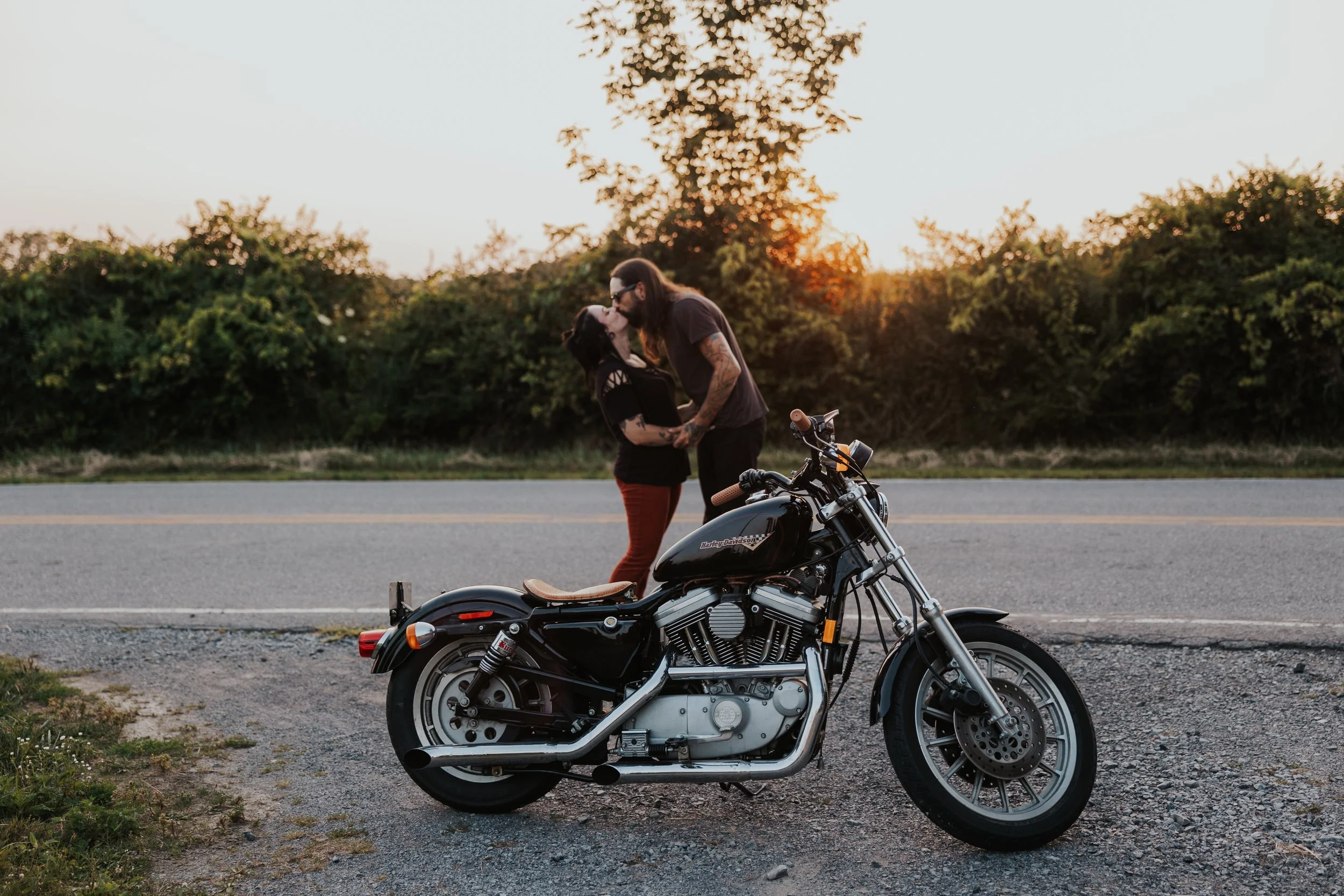 A couple kissing next to a black Harley-Davidson motorcycle on a roadside during sunset, with trees in the background.