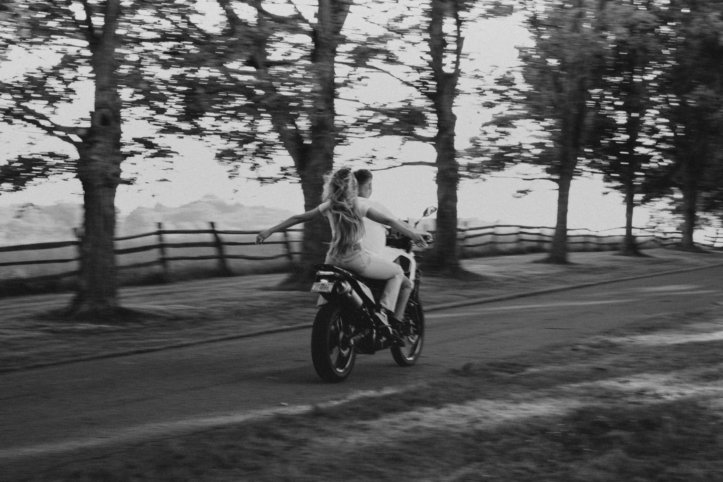 Black and white photo of a woman and a young girl riding a motorcycle along a tree-lined path.