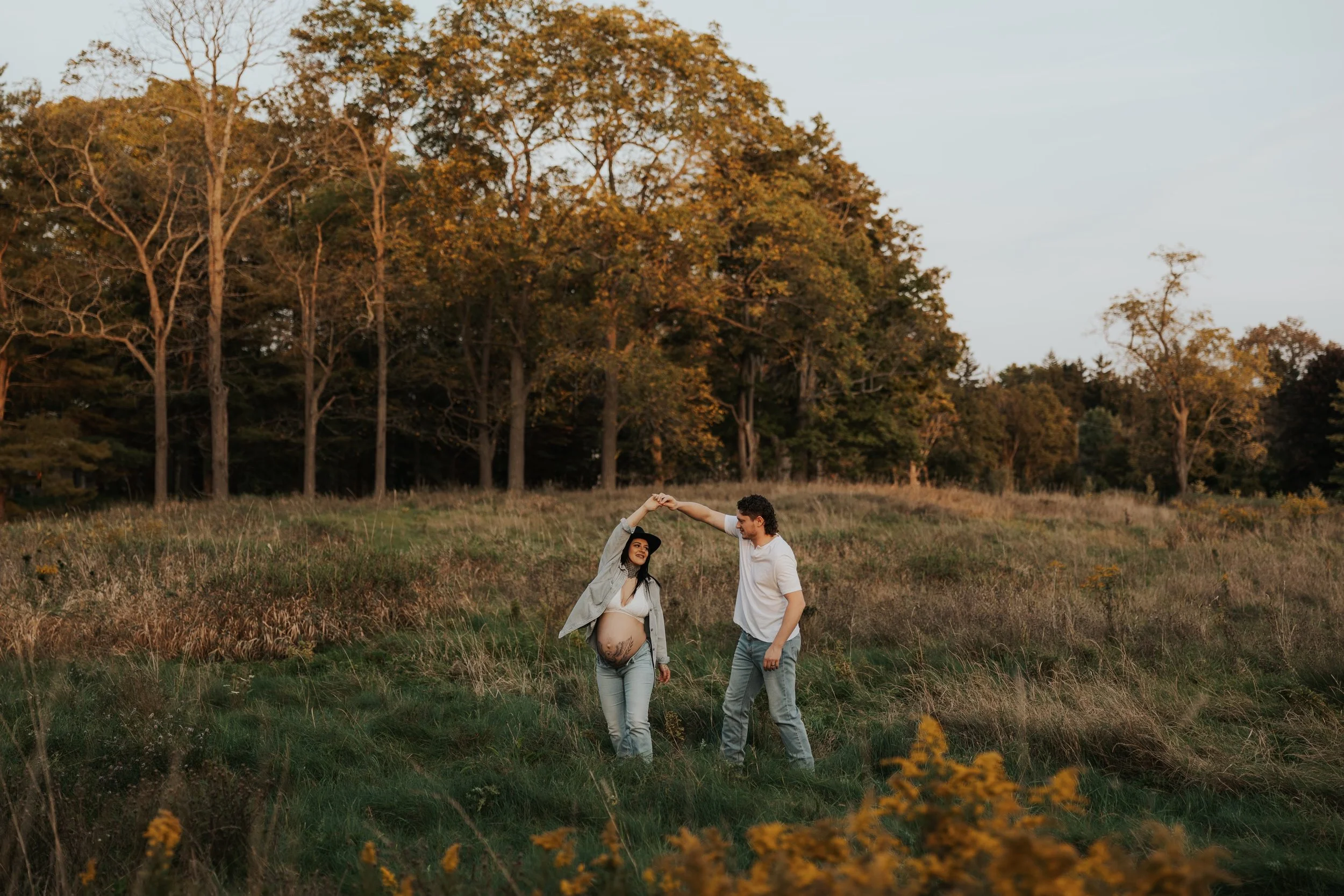 A pregnant woman and a man dancing outdoors in a grassy field with trees in the background during sunset.