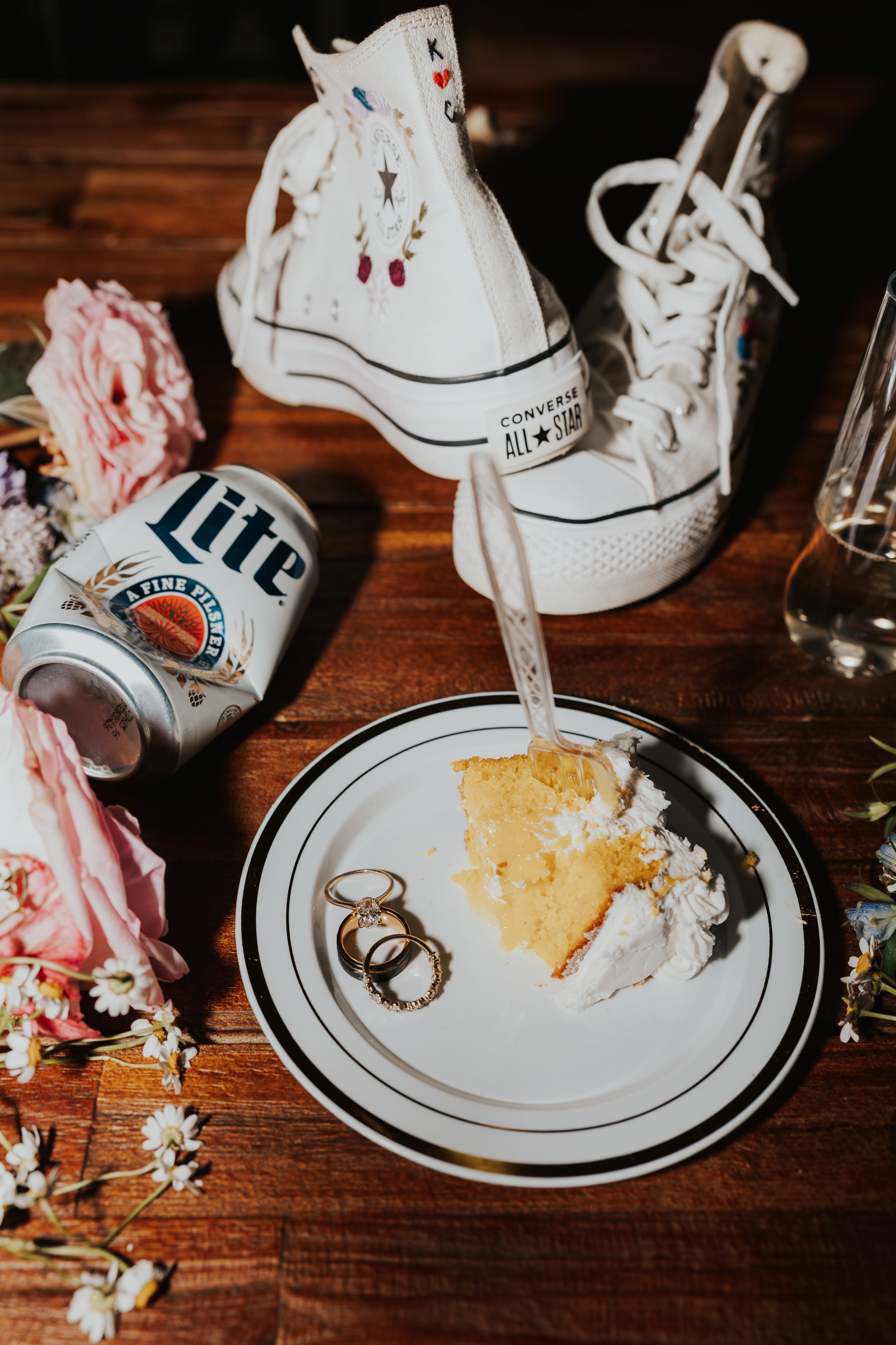 Slice of vanilla cake with whipped cream on a black and white striped plate, two rings on the plate, a partially eaten cake fork, a can of Miller Lite beer, a pair of white Converse All-Star sneakers, and flowers on a wooden table.