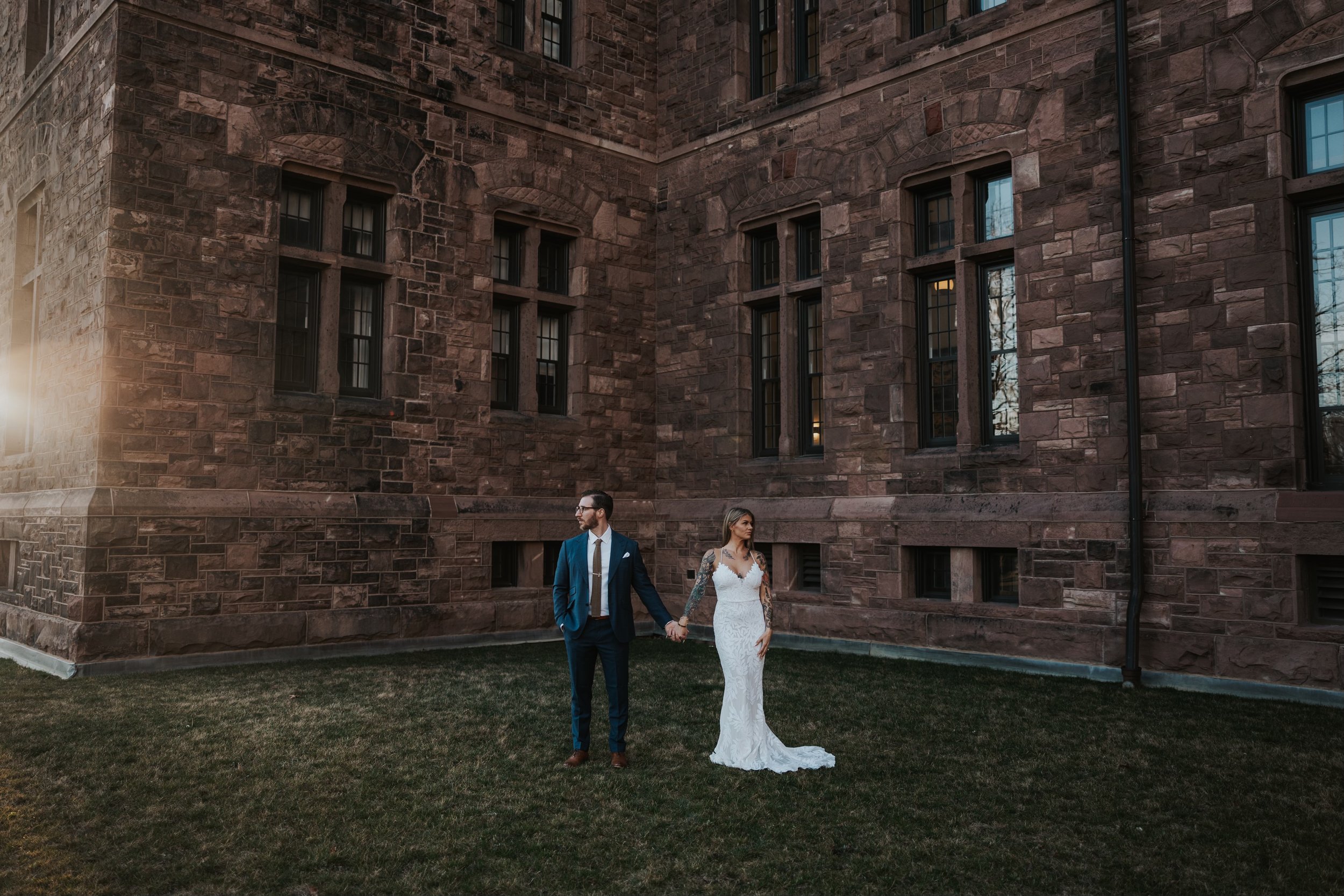 A bride and groom holding hands on a grassy area in front of a historic stone building at sunset