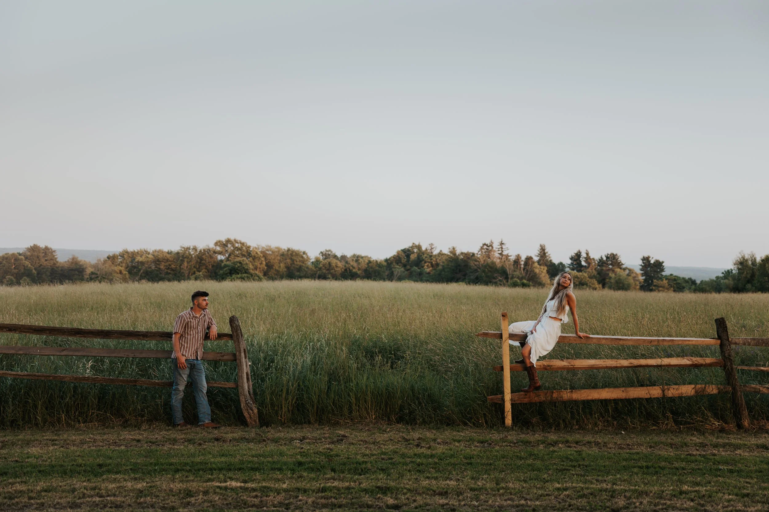 A young man standing near a wooden fence on the left side, looking towards a young woman sitting on the right side of the fence in a field with tall grass and trees in the background during sunset or late afternoon.