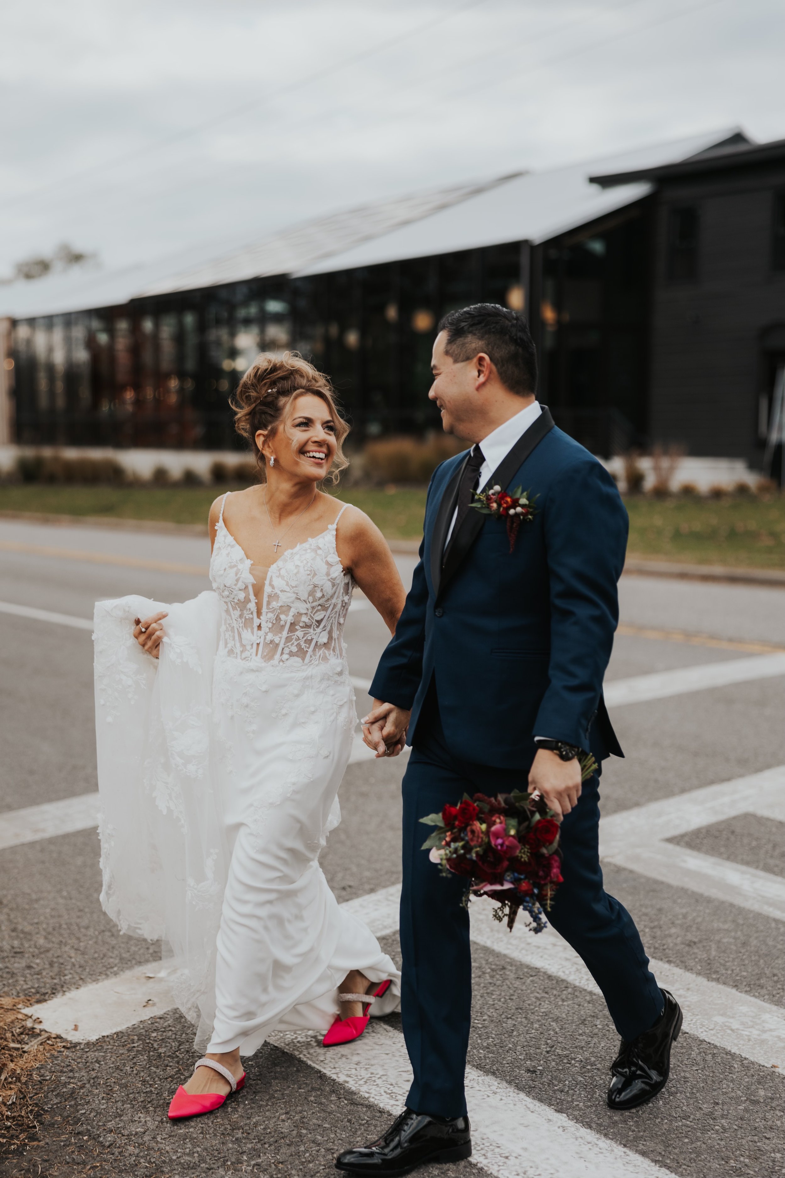 A bride and groom walking hand in hand across a crosswalk on their wedding day, smiling at each other. The bride is in a lace wedding dress with bright pink shoes, and the groom is in a navy suit holding a bouquet of flowers.