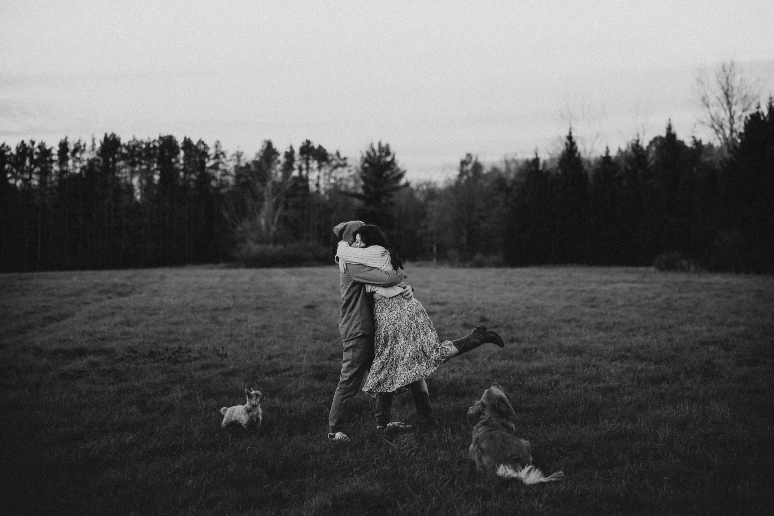 A black-and-white photo of two people hugging in a grassy field, with a woman lifting her leg in excitement, and two dogs sitting nearby. There are trees and a cloudy sky in the background.