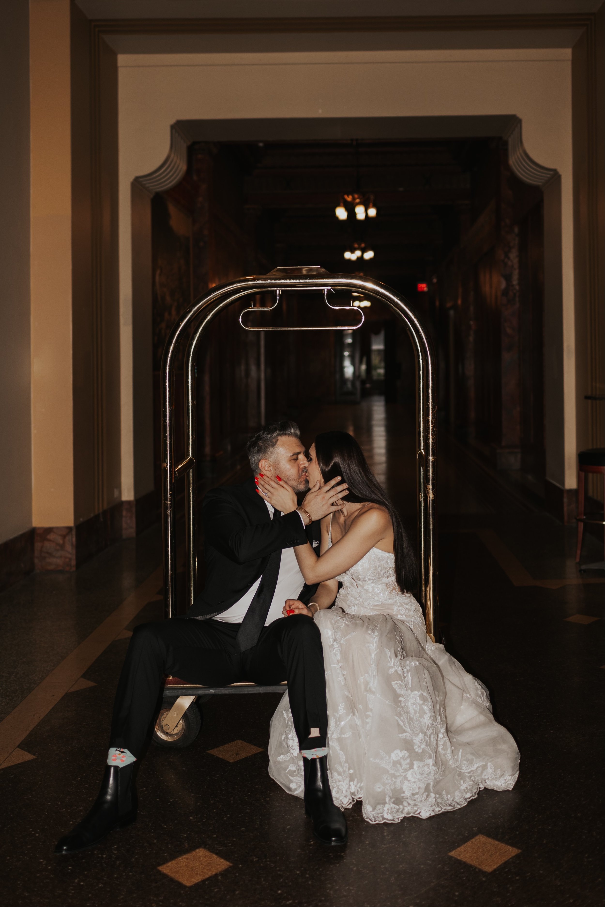 A couple dressed in wedding attire sharing a kiss while sitting on a luggage cart in an indoor hallway.
