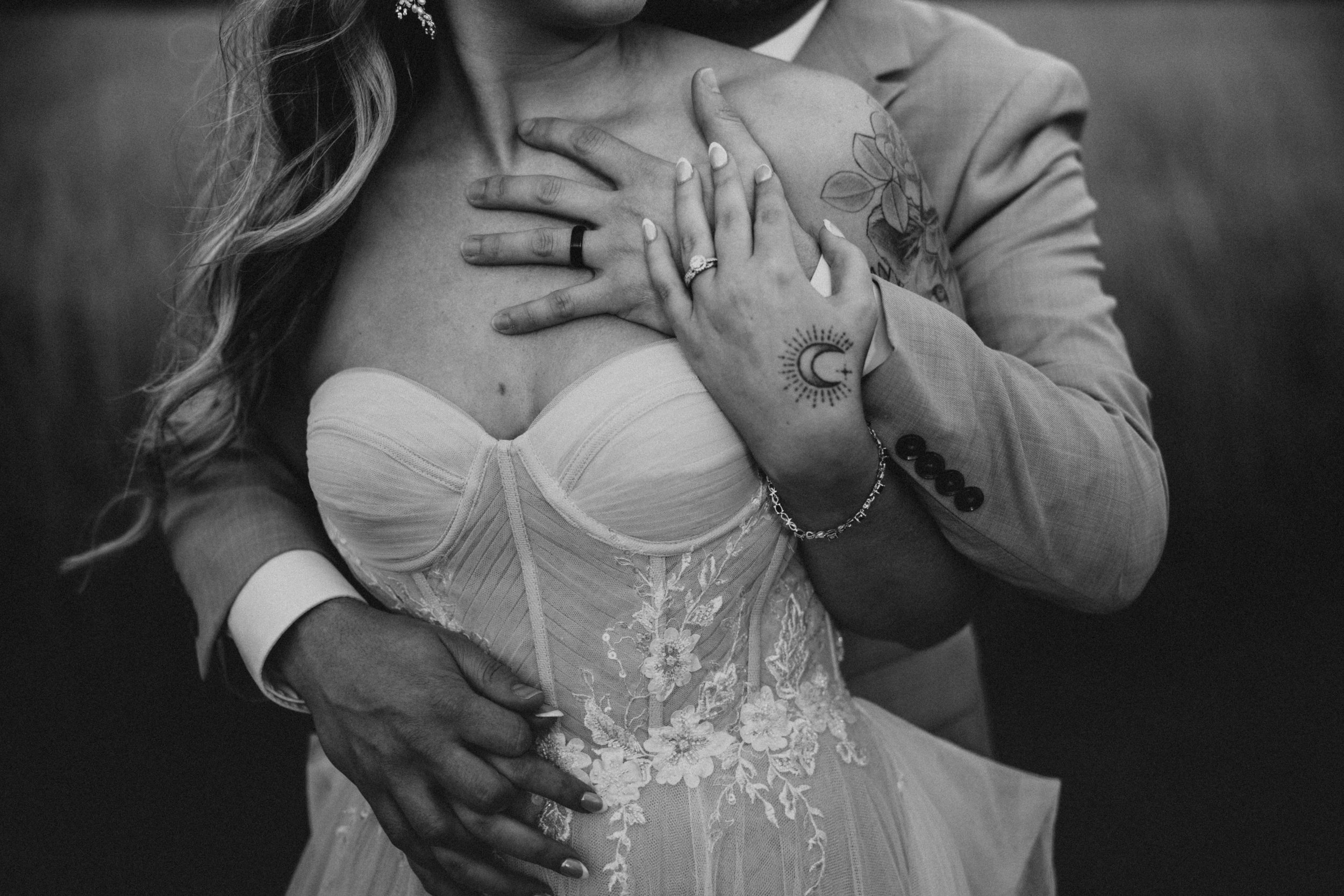 A black and white photo of a couple, with the man embracing and holding the woman, who is wearing a wedding dress. The woman's hand, with an engagement ring, is resting on her chest, and both show tattoos, including a moon and sun design. The man is