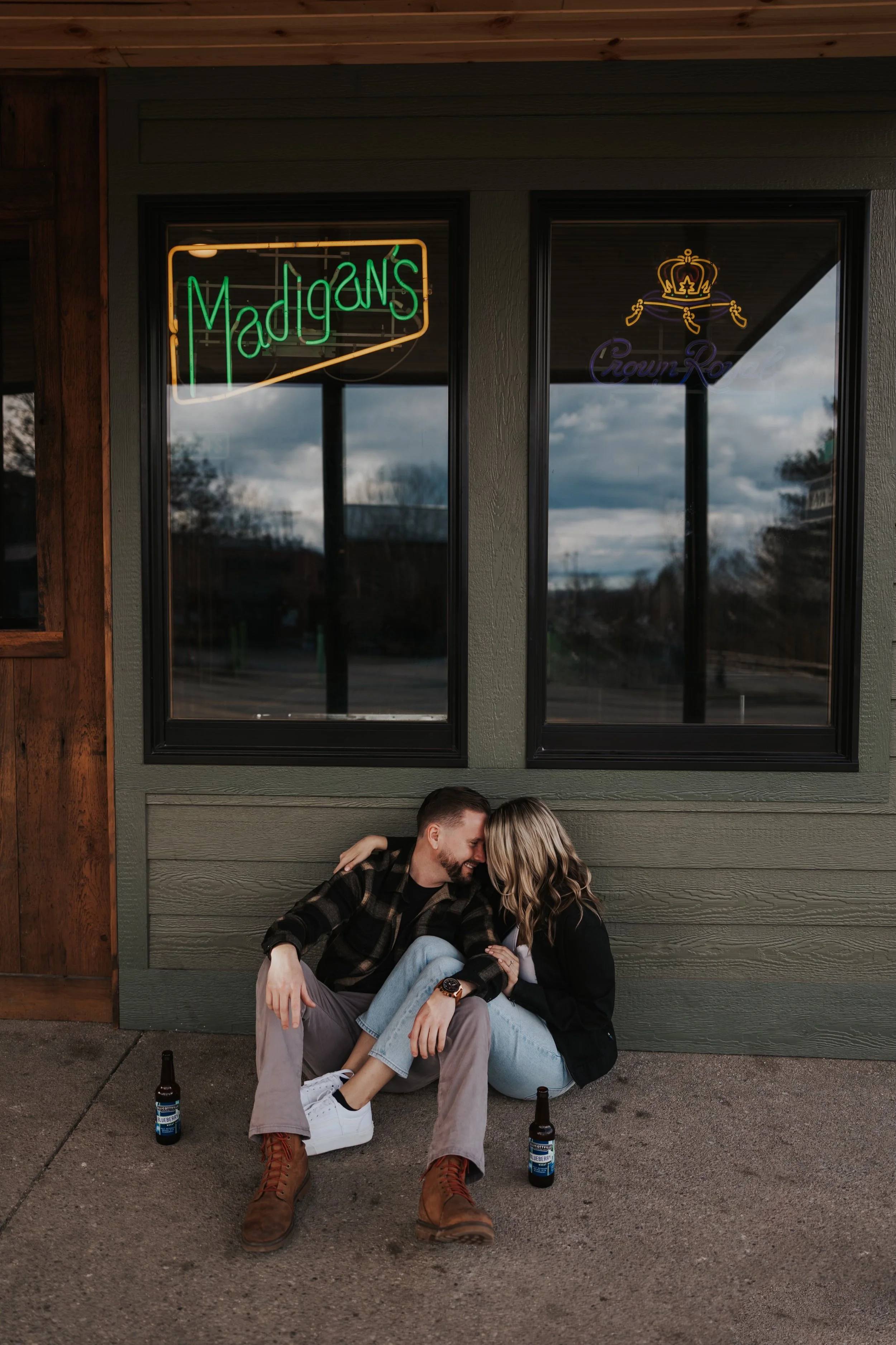 A young couple sitting on the sidewalk outside a restaurant or bar, smiling and leaning into each other, with two beer bottles on the ground. Behind them, there are two large windows with neon signs, one spelling 'Jadgns' and the other featuring a cr