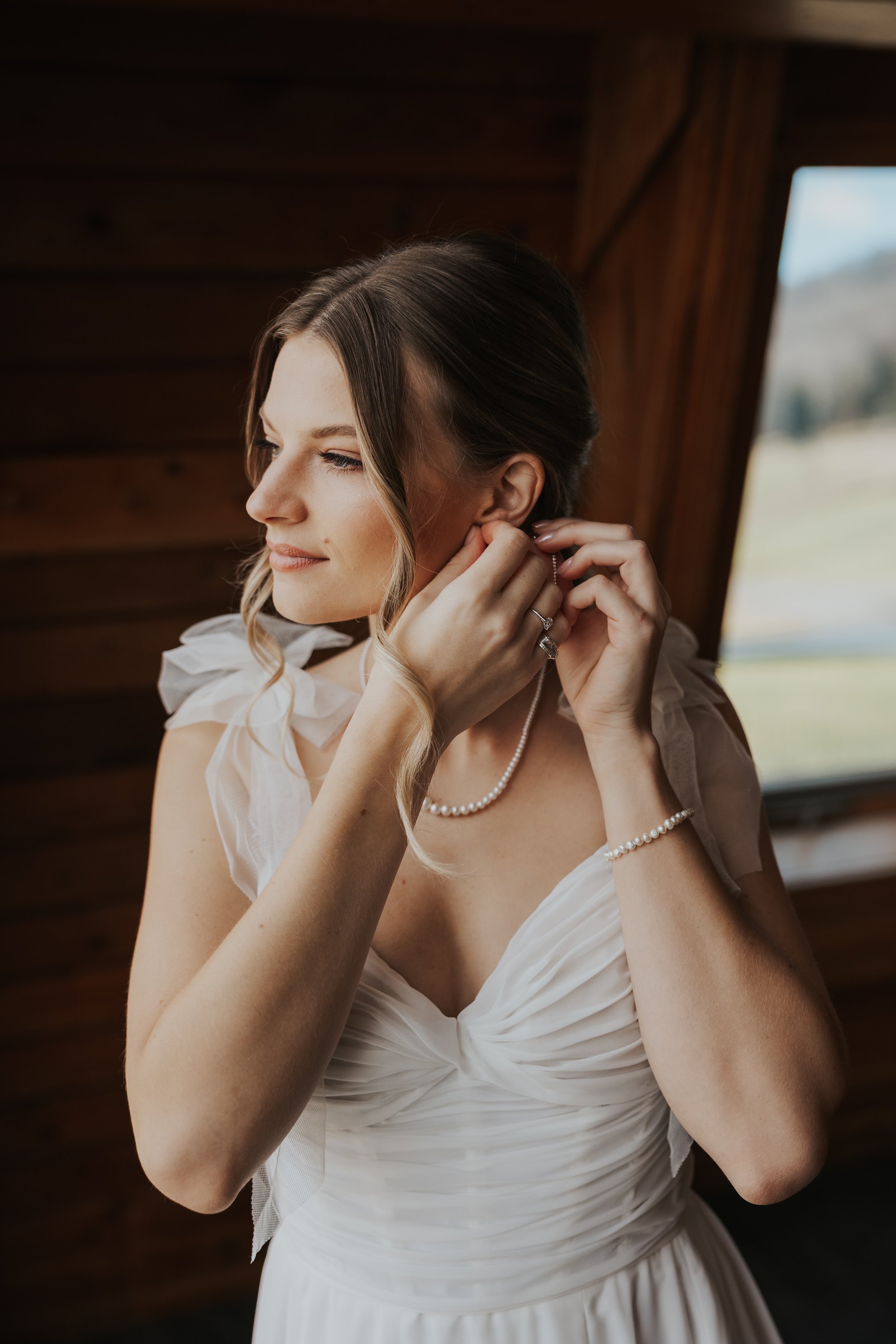 A bride wearing a white wedding dress, pearl necklace, bracelet, and earrings, adjusting her earring inside a wooden room with a window showing a landscape outside.