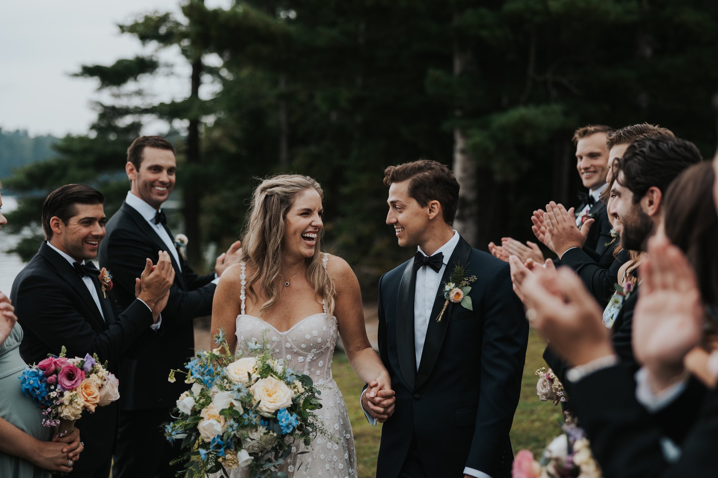 A wedding celebration outdoors with a bride and groom holding hands, surrounded by friends clapping and smiling.
