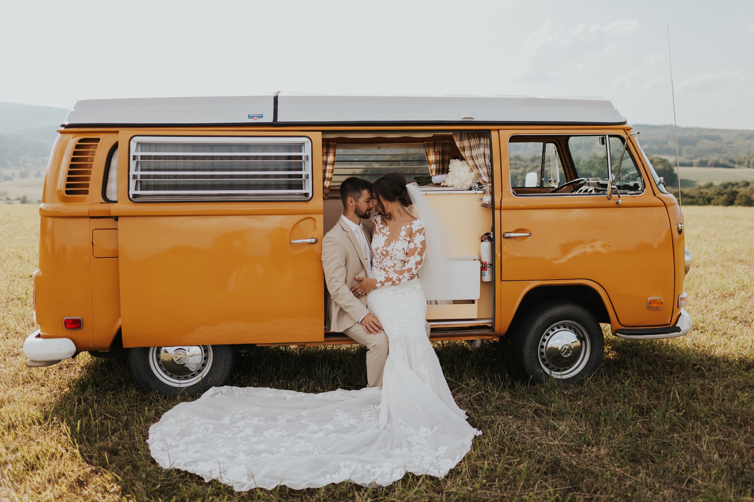 A bride and groom standing in front of a vintage orange camper van on a grassy field during the day, with the bride in a white lace wedding dress and veil, and the groom in a beige suit.