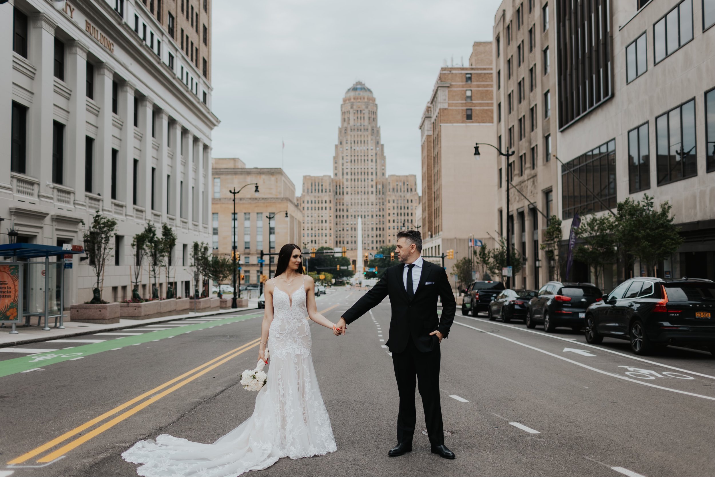 A newlywed couple holding hands in the middle of a city street, with tall buildings and parked cars around them, and a city skyline in the background.