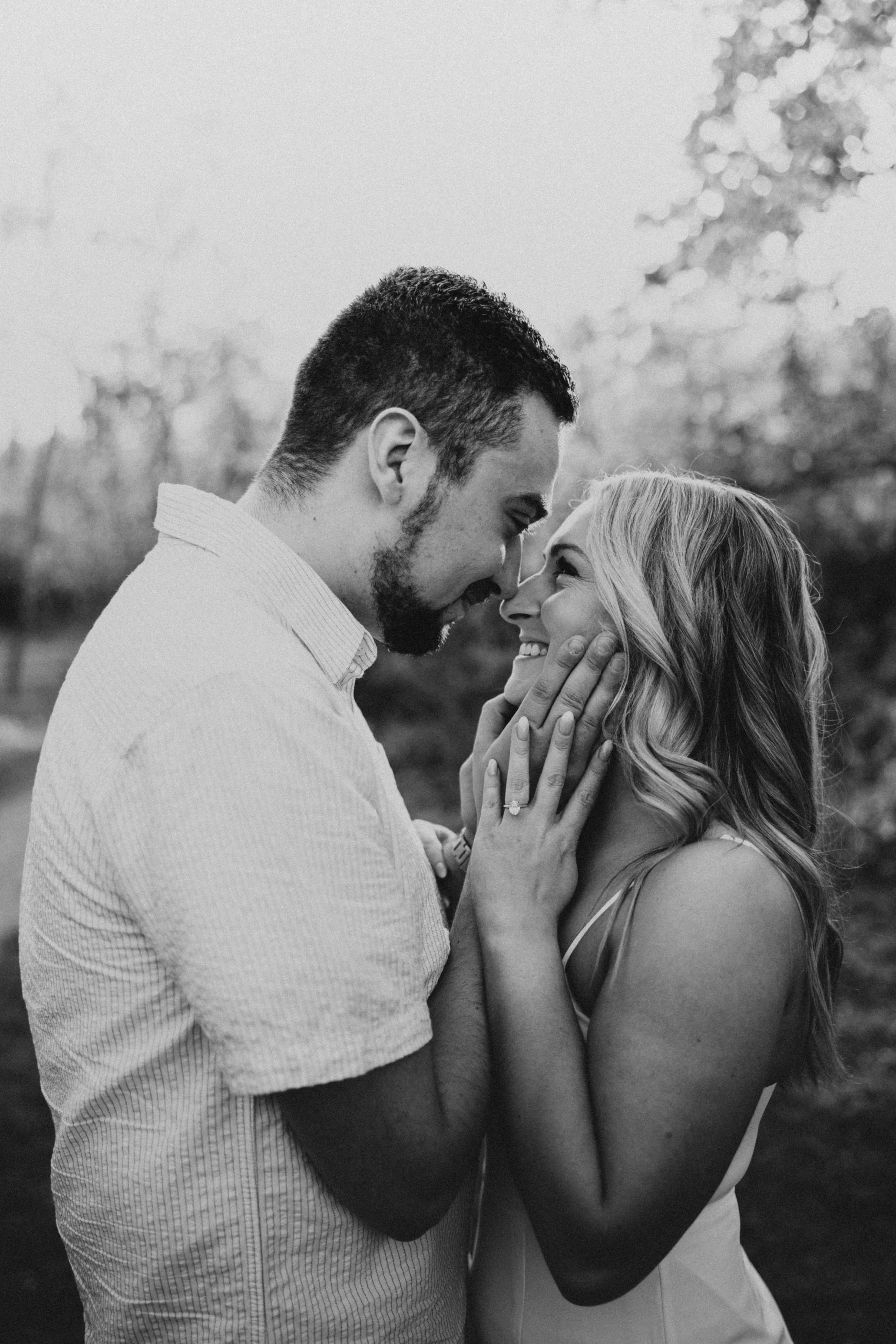 A black and white photograph of a couple sharing an intimate moment outdoors, with the man holding the woman's face as they look into each other's eyes and smile.