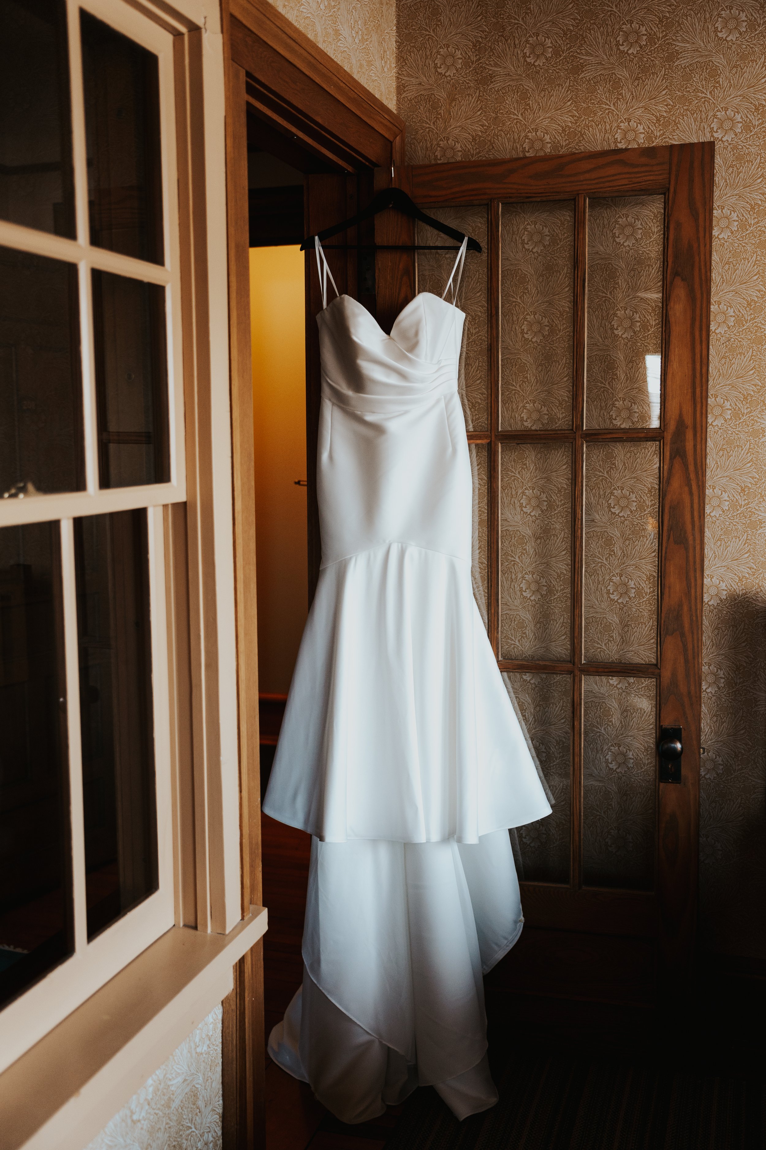 A white wedding dress hangs on a black hanger inside a doorway, with a wooden door and patterned wallpaper in the background.