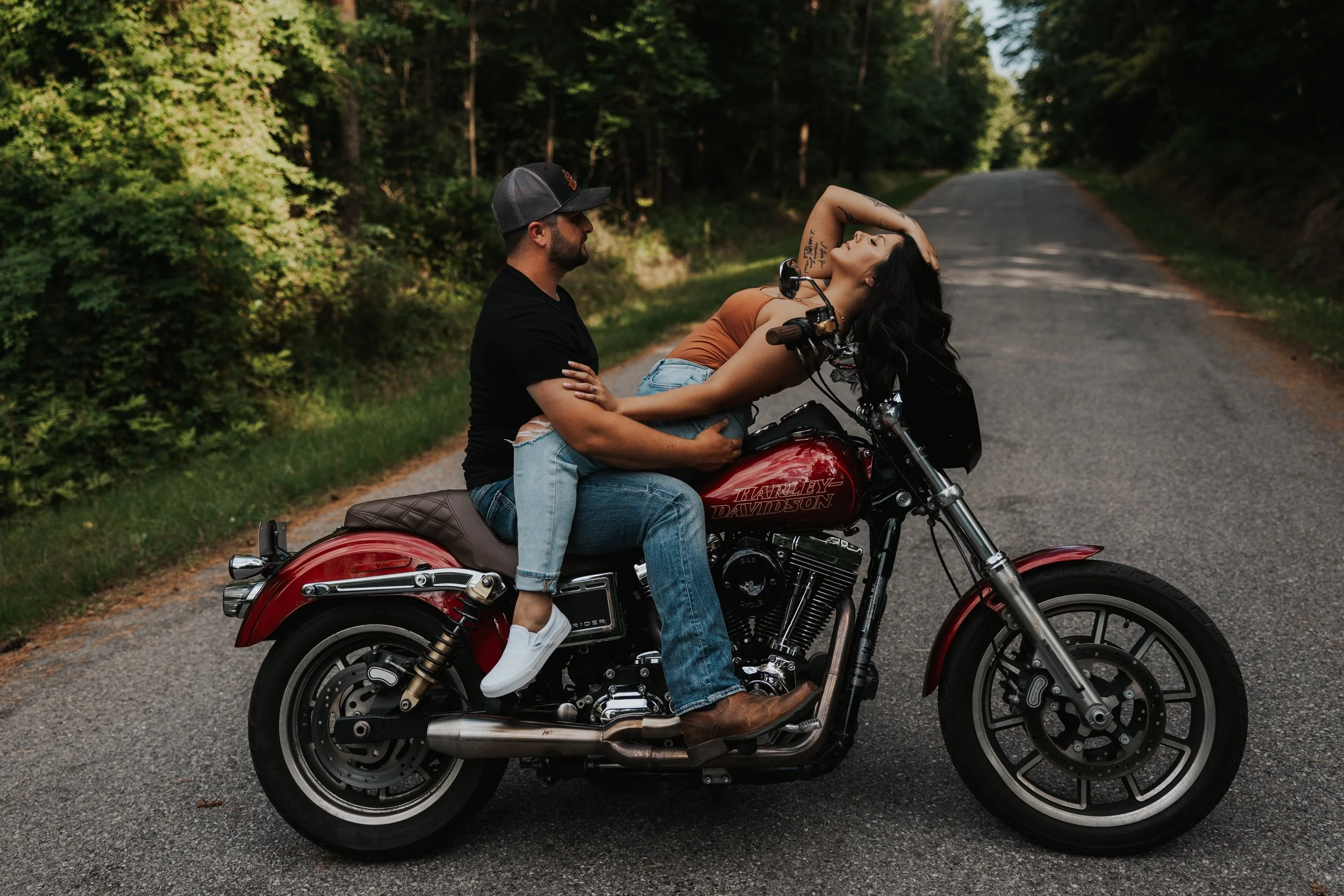 A man and woman are on a red Harley Davidson motorcycle on a rural road, with the woman leaning back and the man holding her, surrounded by green trees.