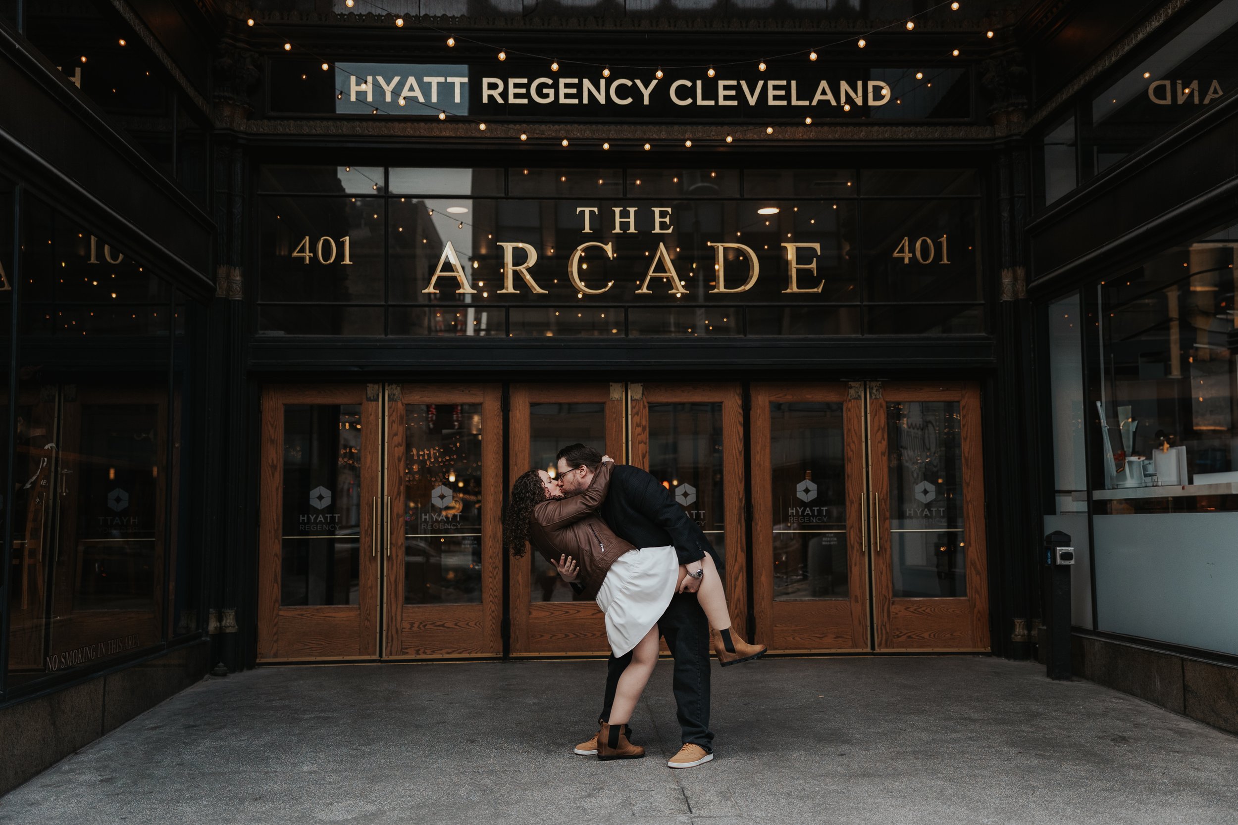A couple sharing a kiss in front of the entrance to The Arcade at Hyatt Regency Cleveland, with wood doors and large glass windows reflecting lights inside.