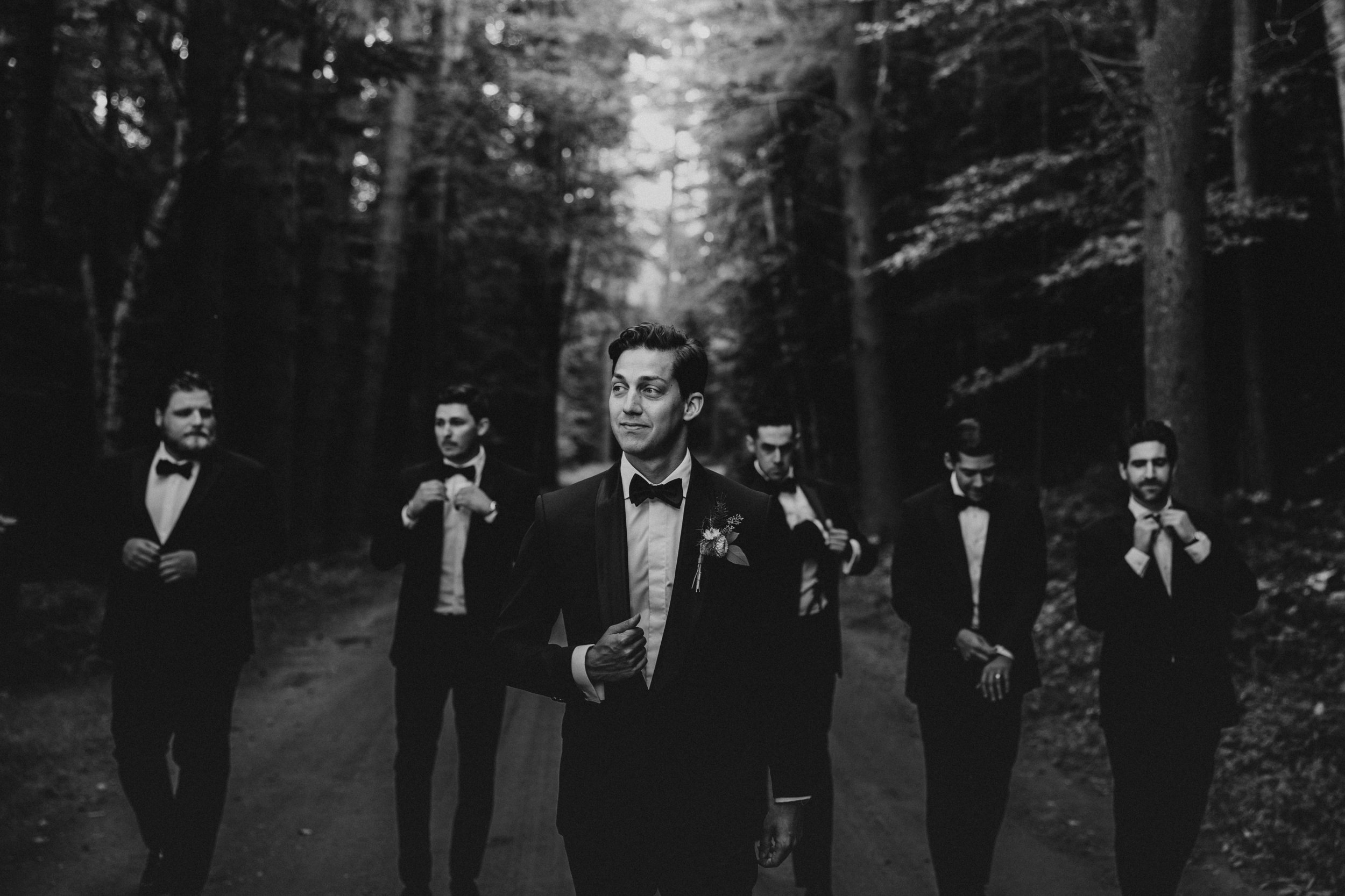 A groom and five groomsmen in tuxedos in a forest, with the groom in front and the others adjusting their bow ties.