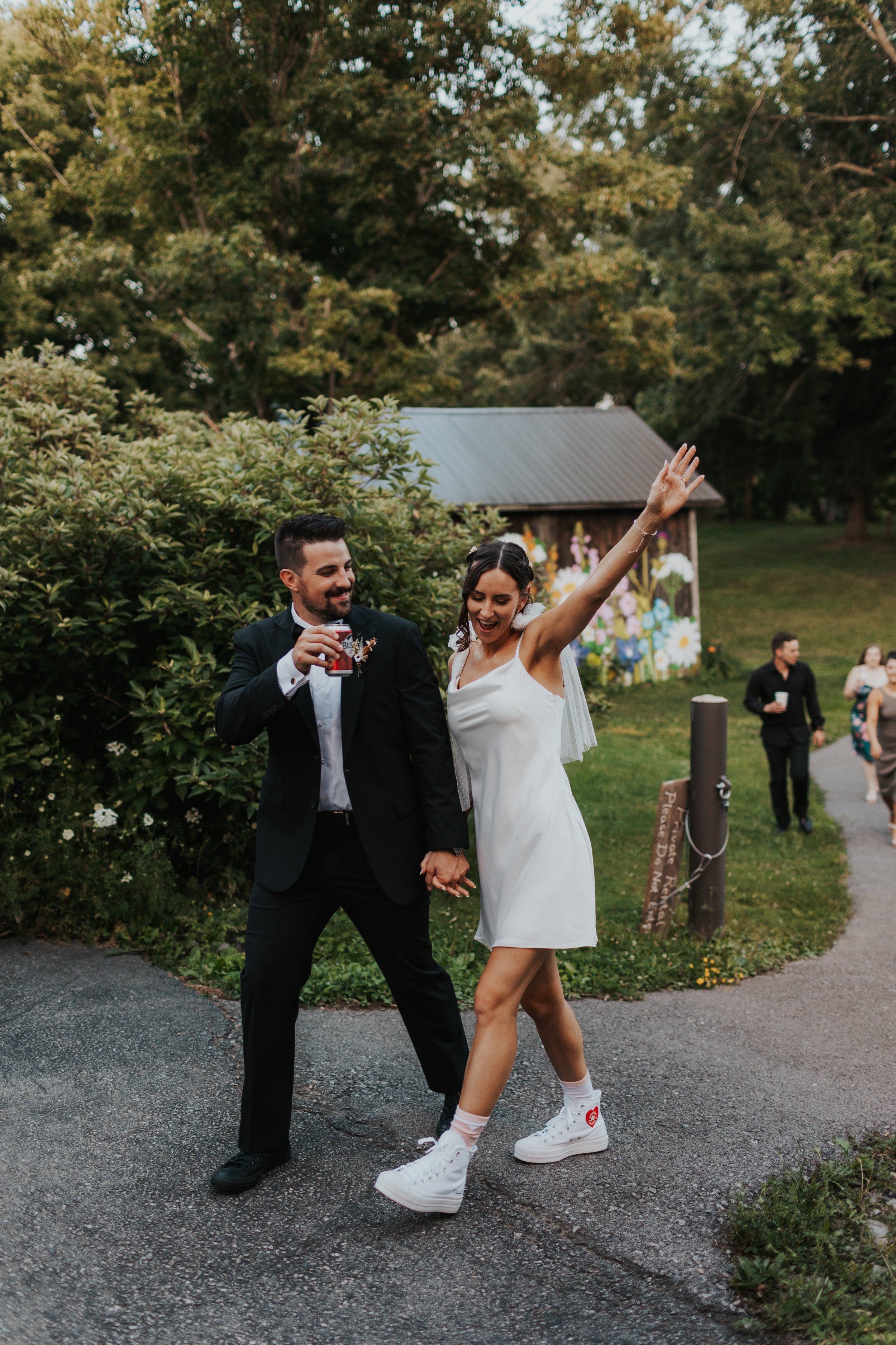 A joyful couple is dancing outdoors at a wedding reception, holding hands and smiling. The woman is wearing a white slip dress and white sneakers, with her arm raised, and the man is dressed in a black suit and white shirt, holding a drink in his oth