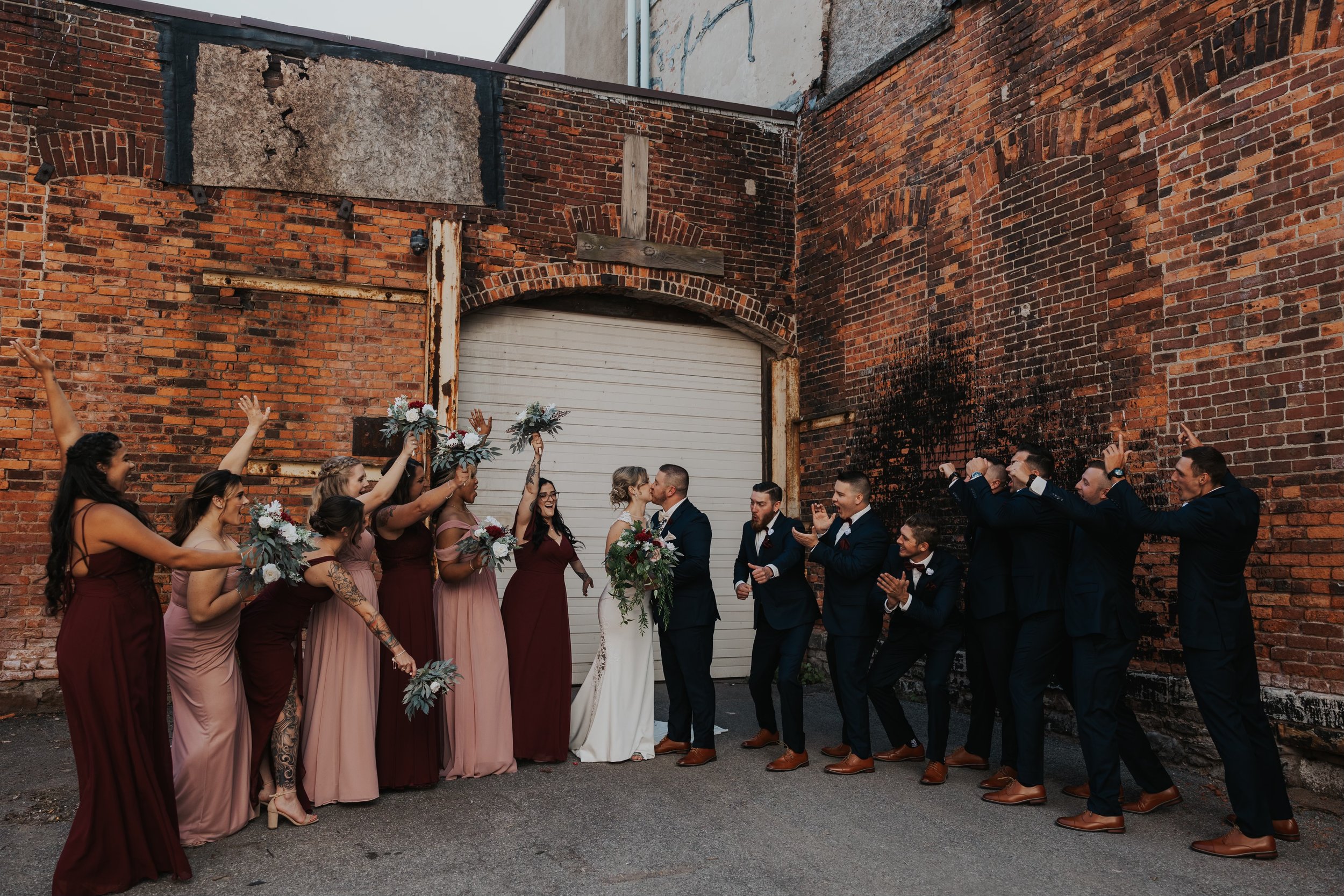 A wedding party celebrating in front of a brick wall, with the bride and groom in the center kissing, surrounded by bridesmaids holding bouquets on the left and groomsmen in suits cheering on the right.