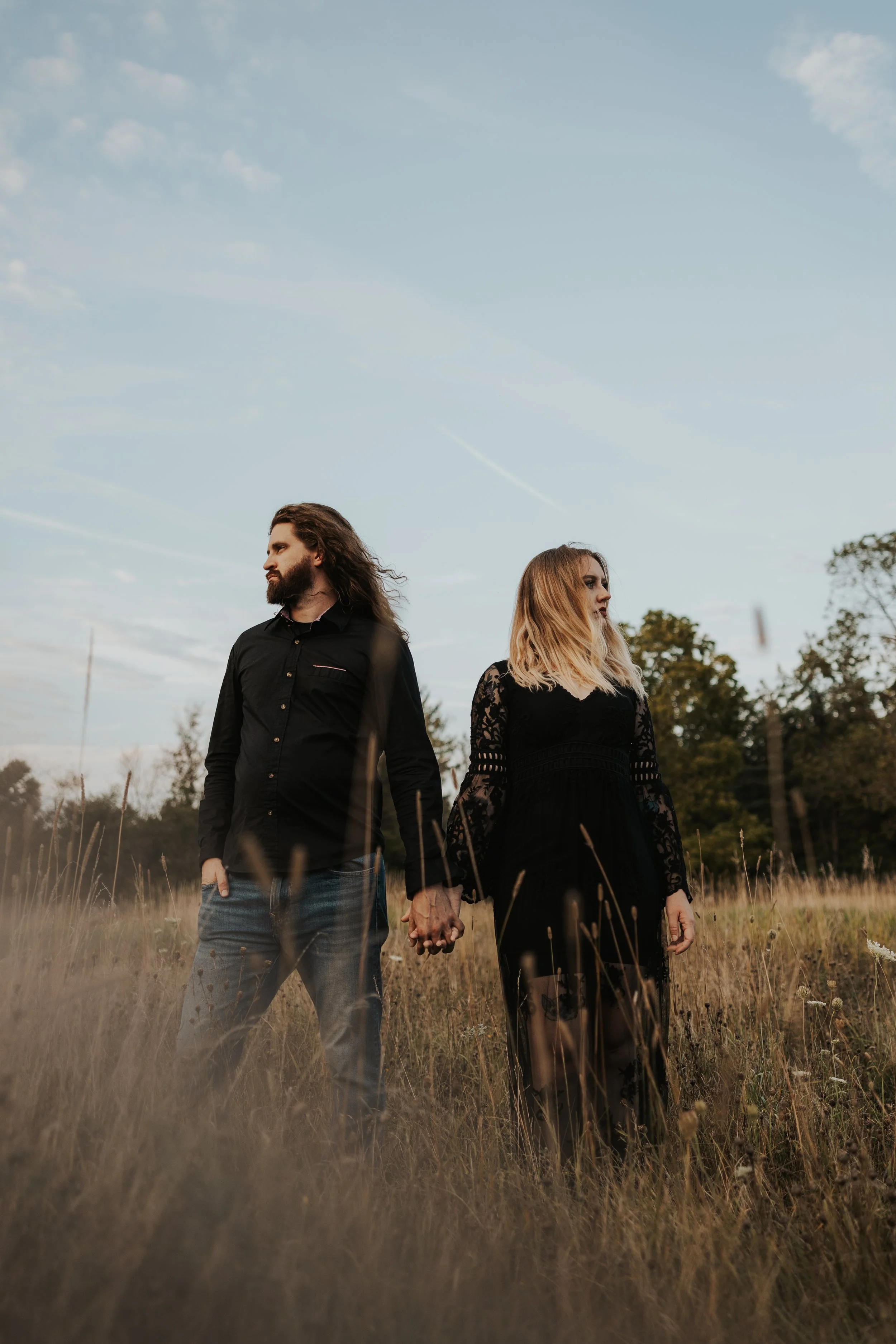 A man and woman holding hands in a field during sunset, both looking away from each other, with the man having long brown hair and beard, wearing a black shirt, and the woman having blonde hair, wearing a black lace dress.