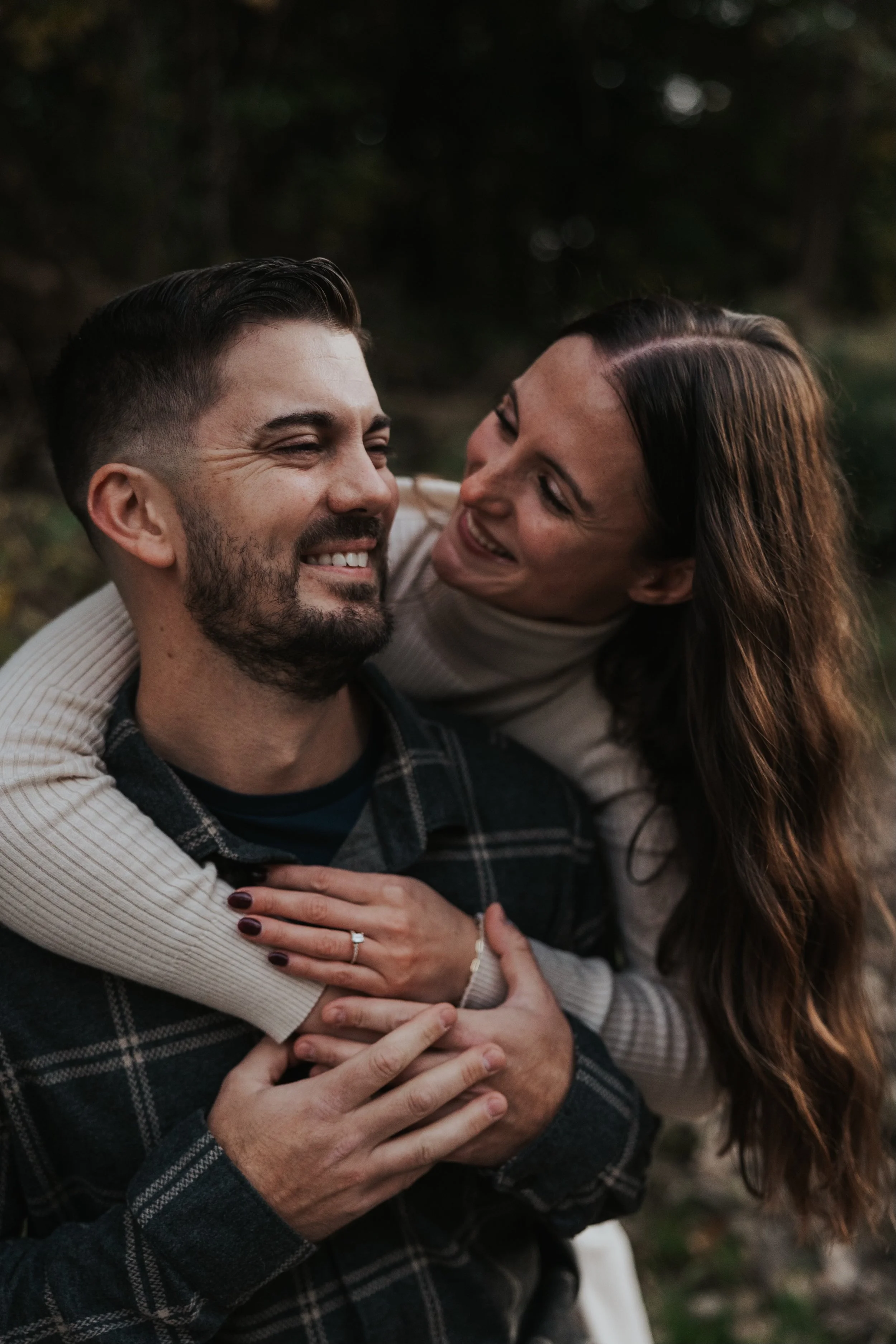 A smiling woman with long brown hair and a man with short dark hair and a beard are outdoors, with the woman embracing the man from behind, both appearing happy and affectionate.