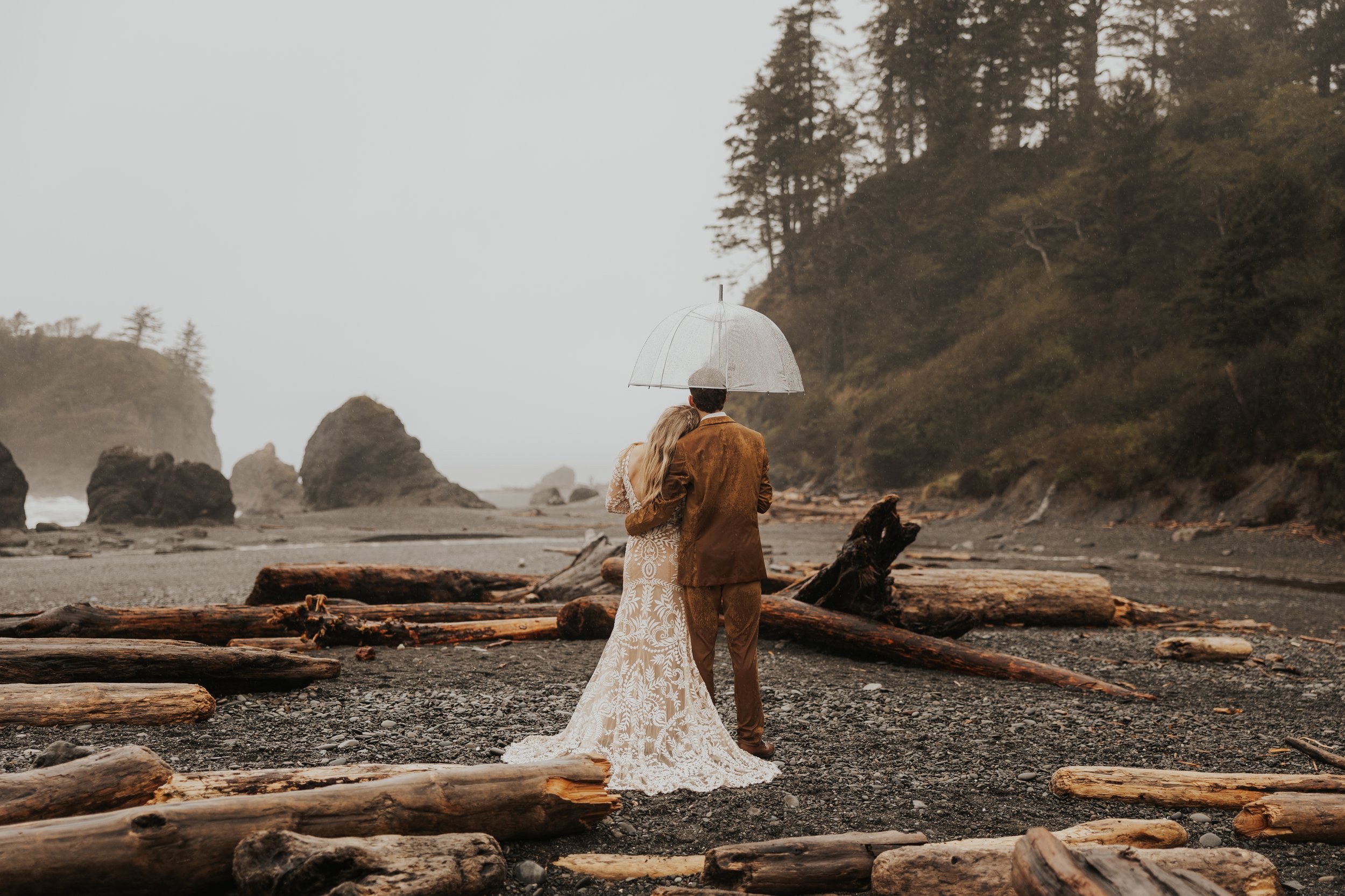 A couple in wedding attire standing on a driftwood-covered beach with a man holding an umbrella over a woman, facing the ocean on a cloudy, foggy day, with forested cliffs in the background.