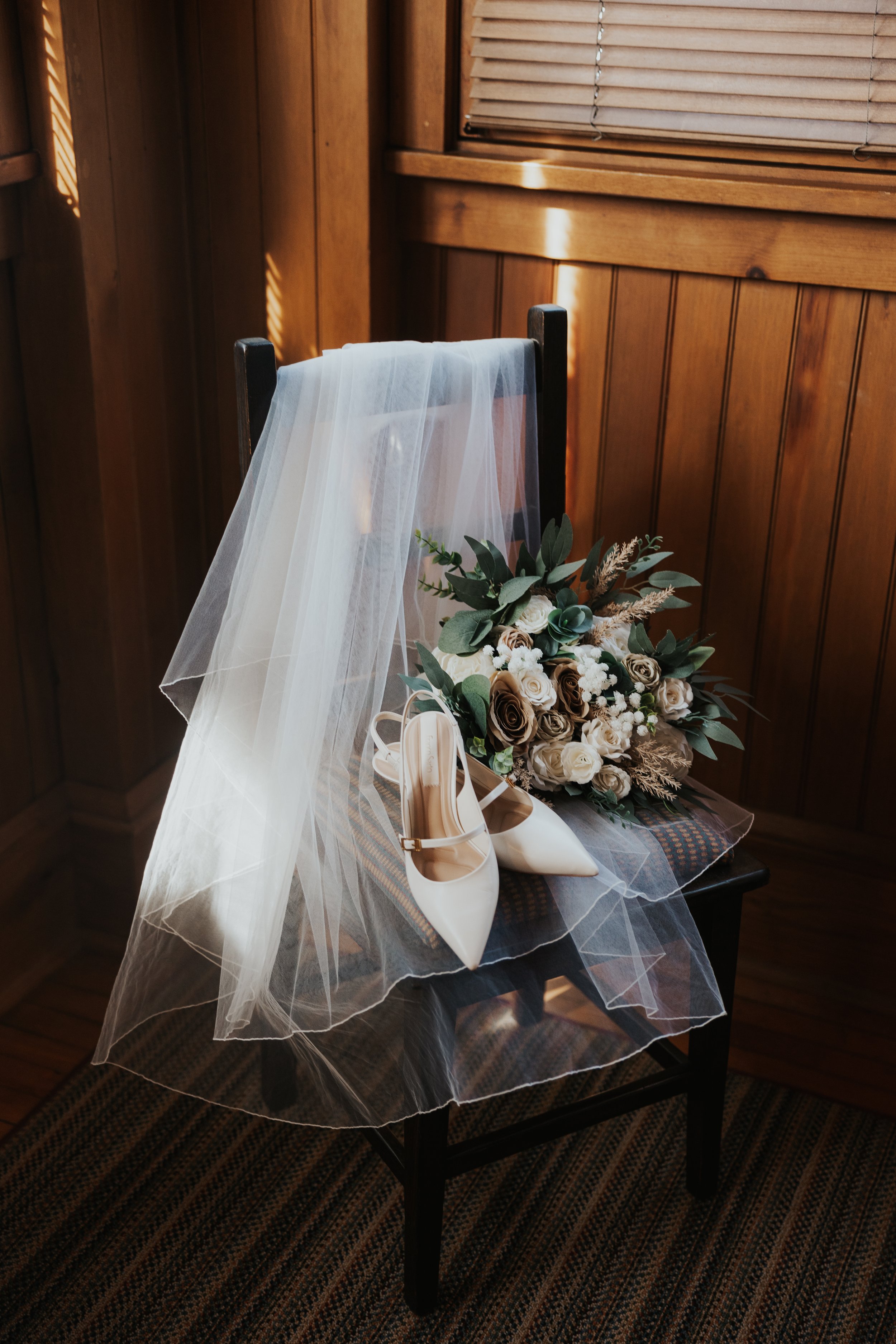 A wooden chair with a white veil draped over the back, a pair of white high-heeled shoes, and a bouquet of white, cream, and taupe roses with greenery on the seat.