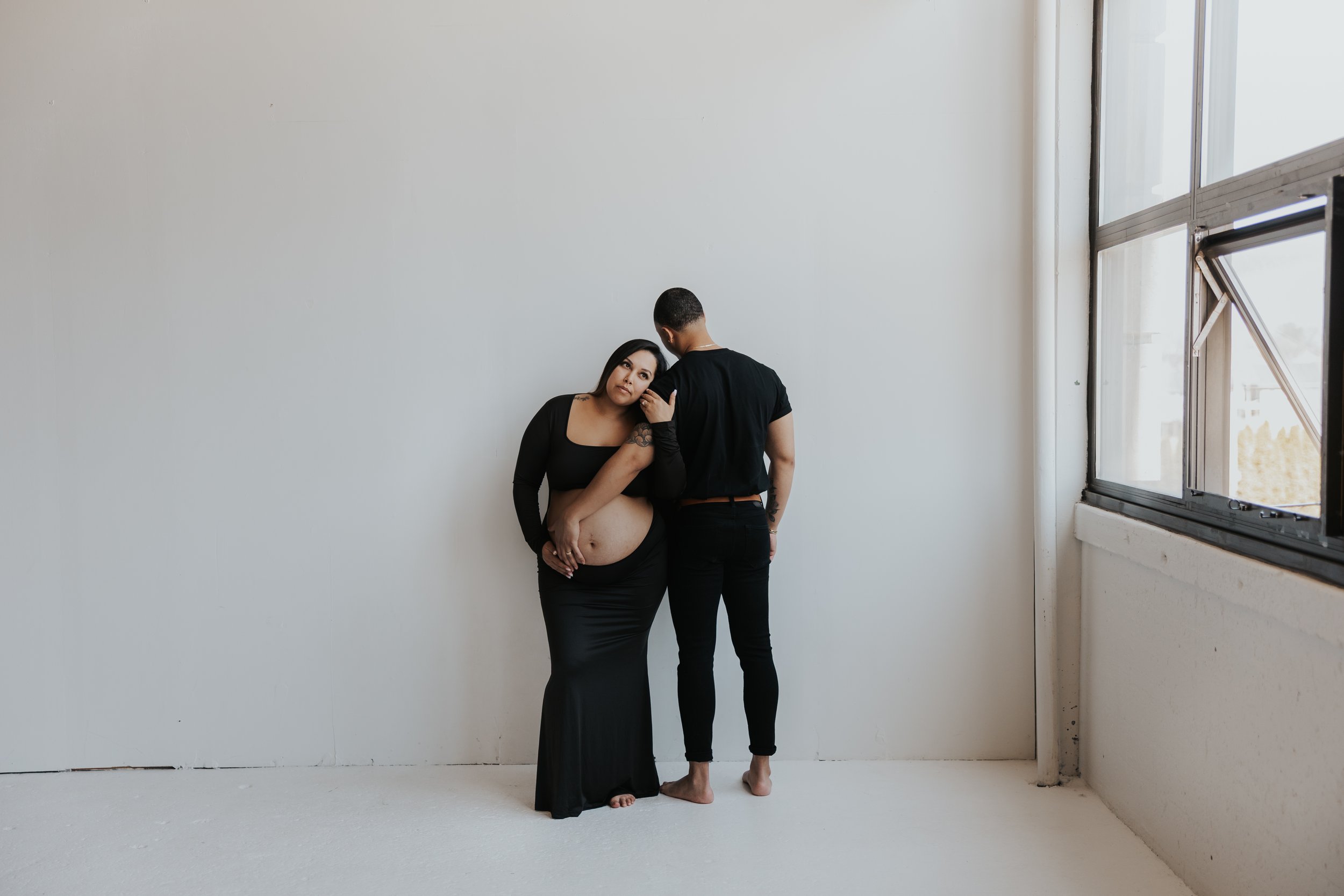 A pregnant woman and her partner standing against a plain white wall near a large window, with the woman touching her belly and looking at the camera, while the man has his back turned.